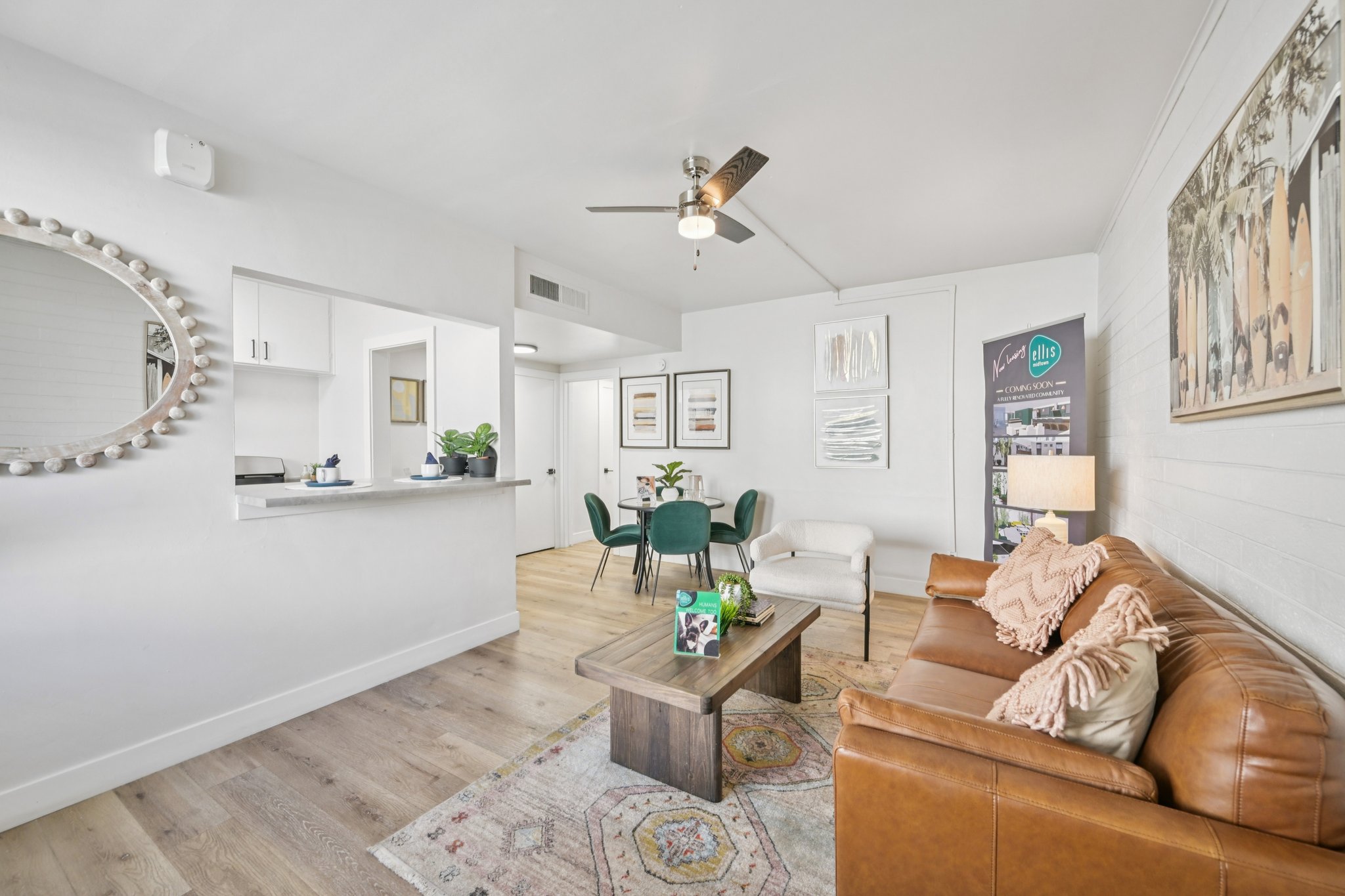 A modern living room featuring a brown leather sofa with decorative pillows, a wooden coffee table on a patterned rug, and a circular mirror on the wall. The space includes a dining area with a round table and green chairs, and the room is well-lit with natural light from a window.