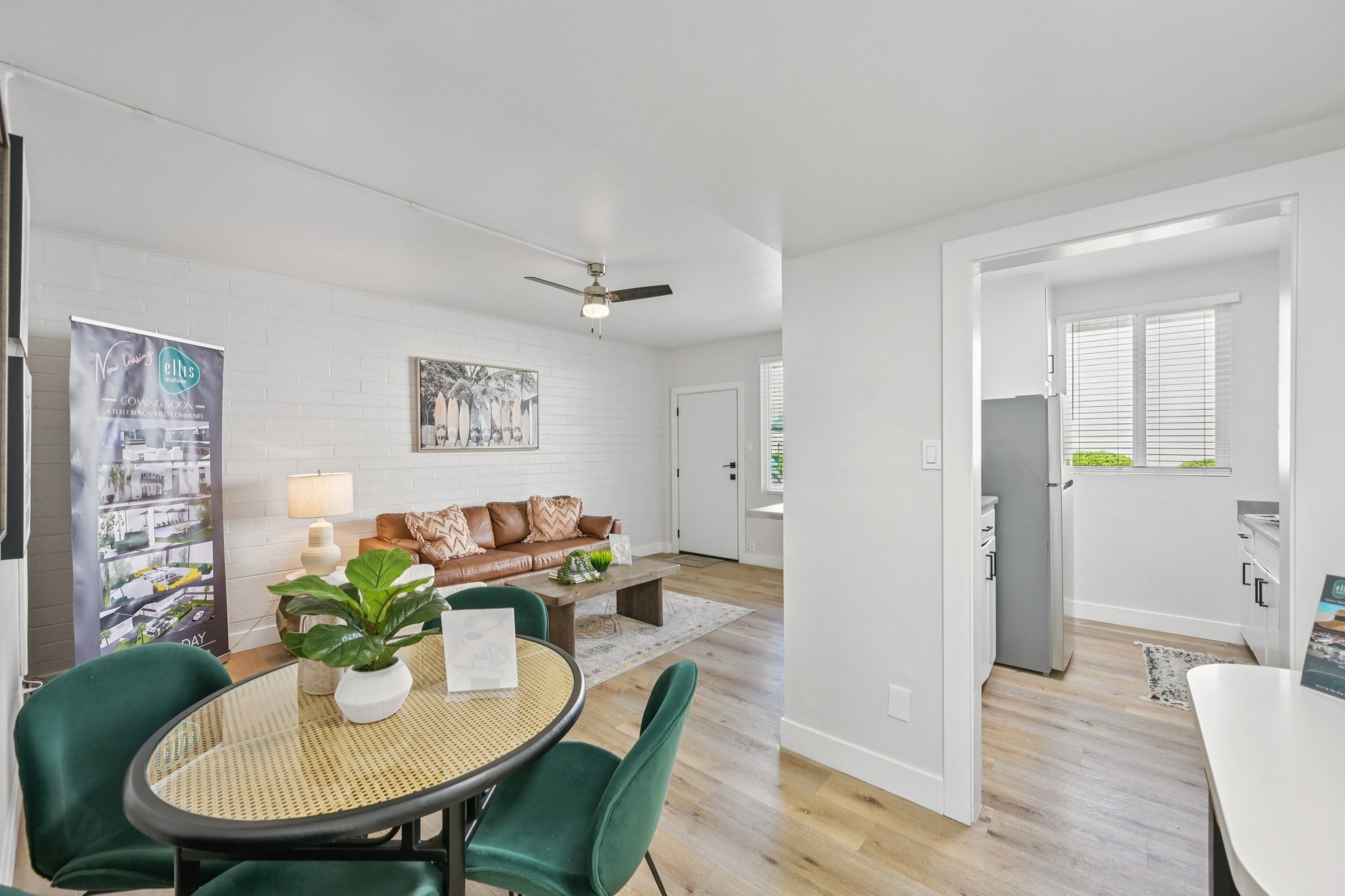 Bright and modern living space featuring a cozy brown leather couch, a round dining table with green chairs, and a decorative plant. Large windows allow natural light to fill the room, with a view into a kitchen area. A banner can be seen in the corner, adding a touch of sophistication to the decor.