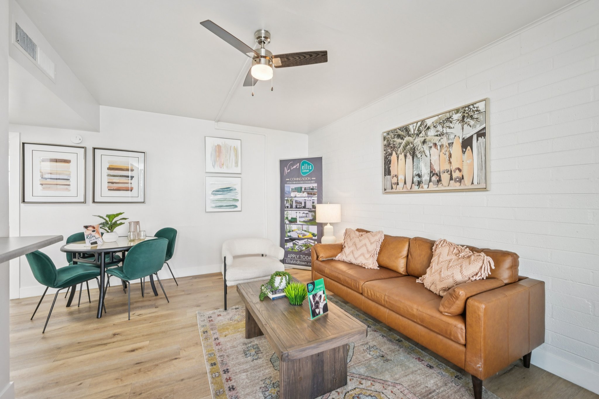 A modern living room featuring a brown leather sofa, a wooden coffee table with decor items, and a ceiling fan. There is a small dining area with green chairs and a round table. Artwork adorns the walls, and a promotional banner is visible. The flooring is light wood, creating a bright and inviting atmosphere.