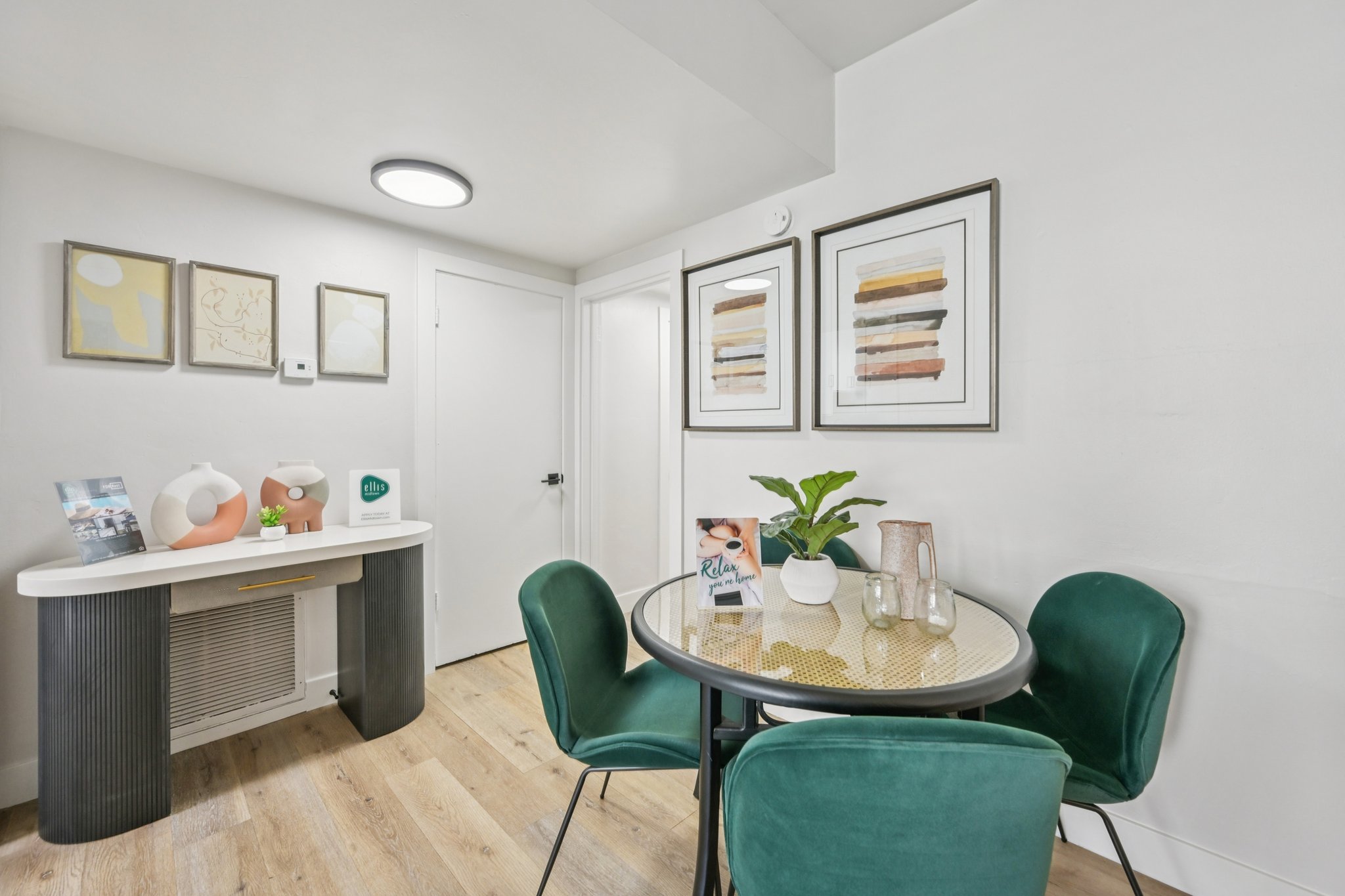 A small, modern dining area featuring a round table surrounded by teal chairs. There is a stylish console table against the wall with decorative items, including a plant and framed artwork above it. The walls are painted white, and the floor is light wood, creating a bright and inviting atmosphere.