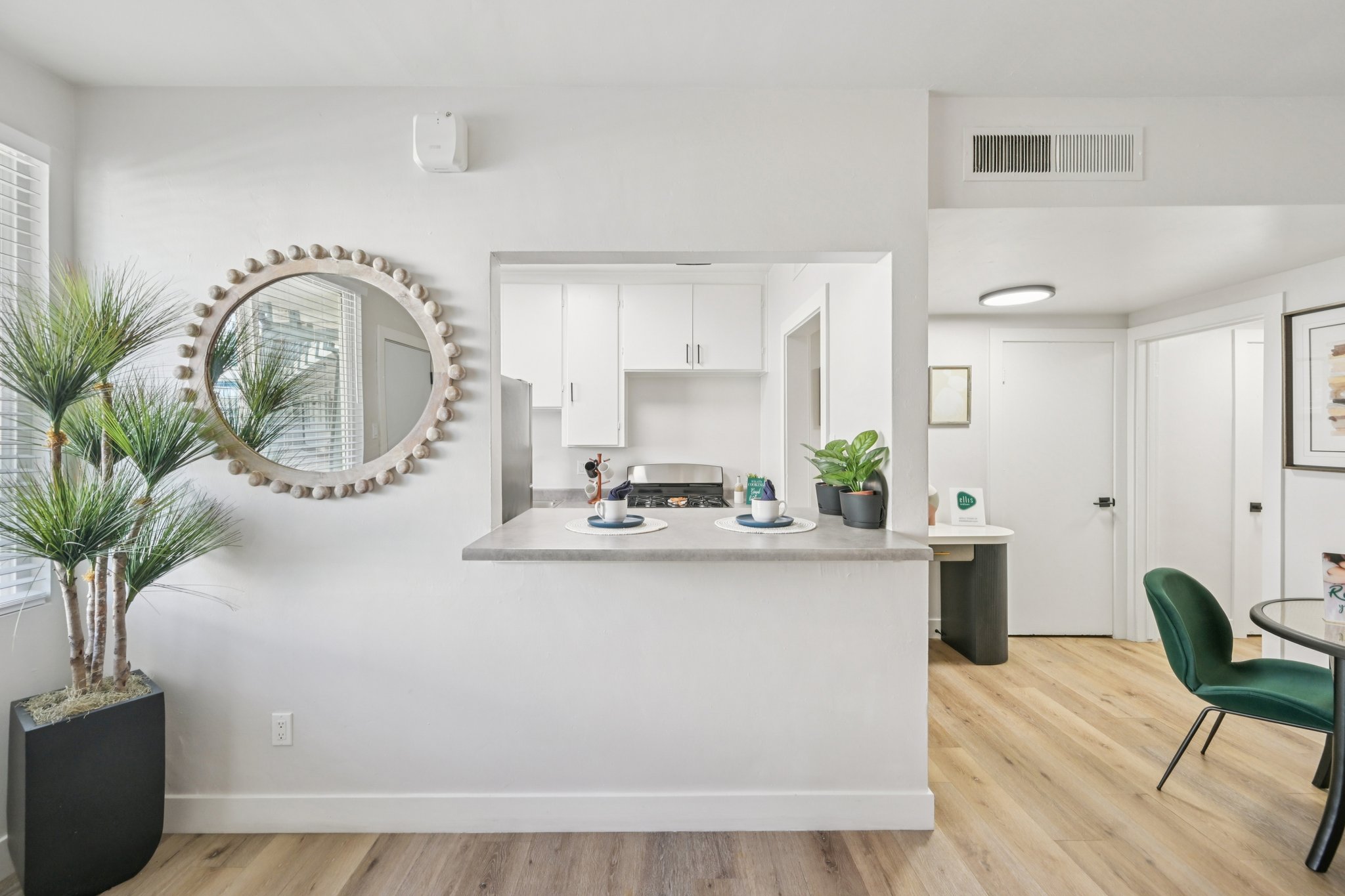 Bright and modern kitchen area featuring a bar counter with two plates and drinks. A decorative round mirror hangs above the counter, surrounded by green plants in stylish pots. Light hardwood flooring complements the neutral walls, while a small dining table with green chairs is visible in the background.