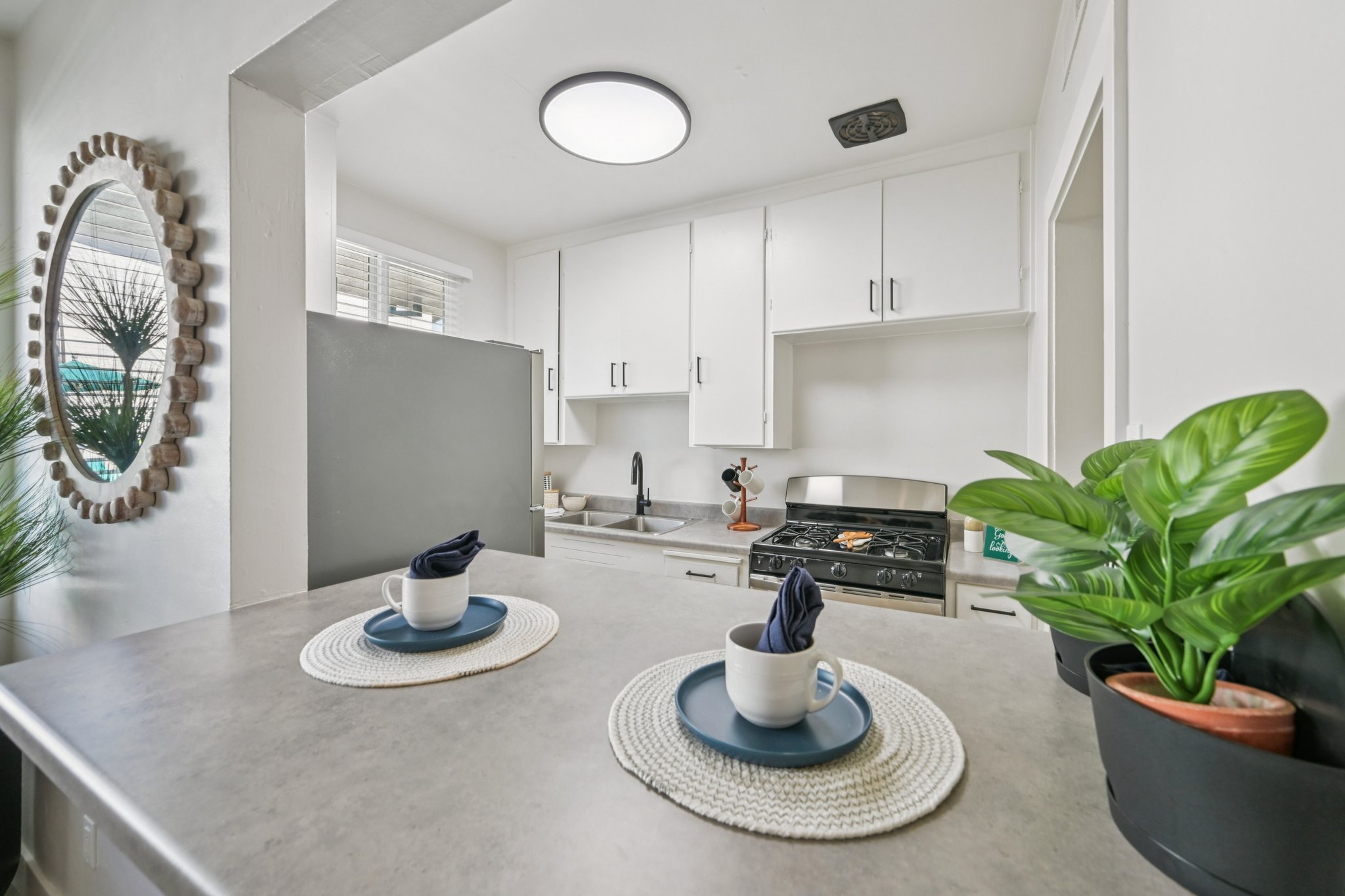 A bright kitchen featuring white cabinets, a modern gas stove, and a sink. A small dining area with two place settings on a gray countertop, including navy napkins and blue plates. There's a round mirror on the wall and a potted plant adding greenery to the space. Natural light enhances the cheerful ambiance.