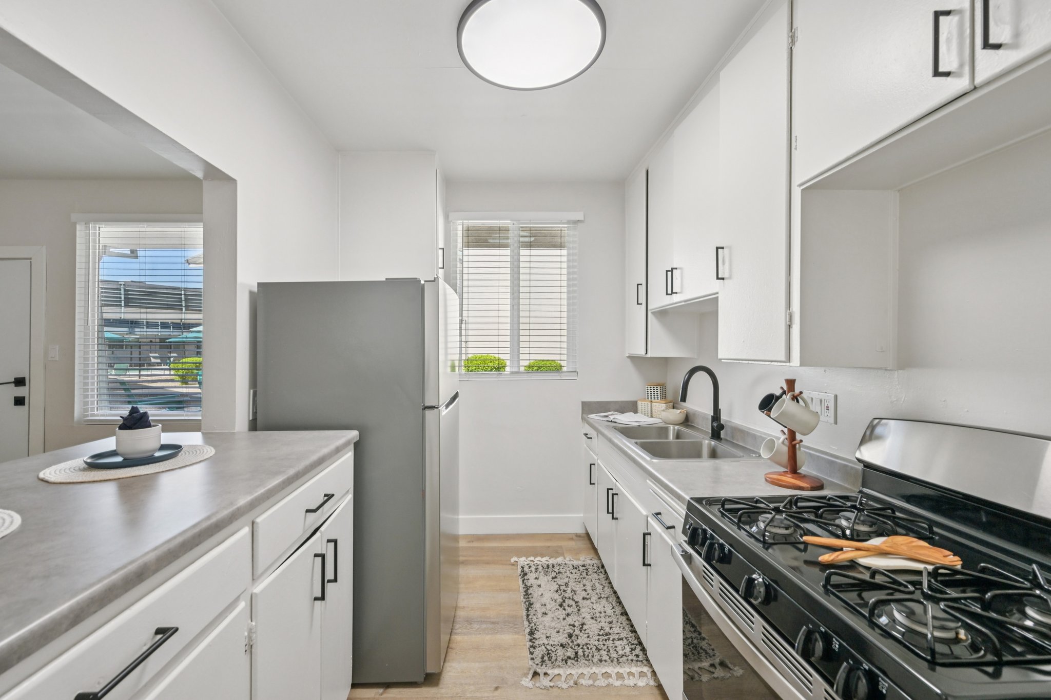 Bright kitchen featuring white cabinetry and a stainless steel refrigerator. A window allows natural light, while a gas stove is positioned next to a stainless sink. Countertop accessories like a wooden cutting board and a decorative plate enhance the space. A patterned rug adds warmth to the wooden flooring.