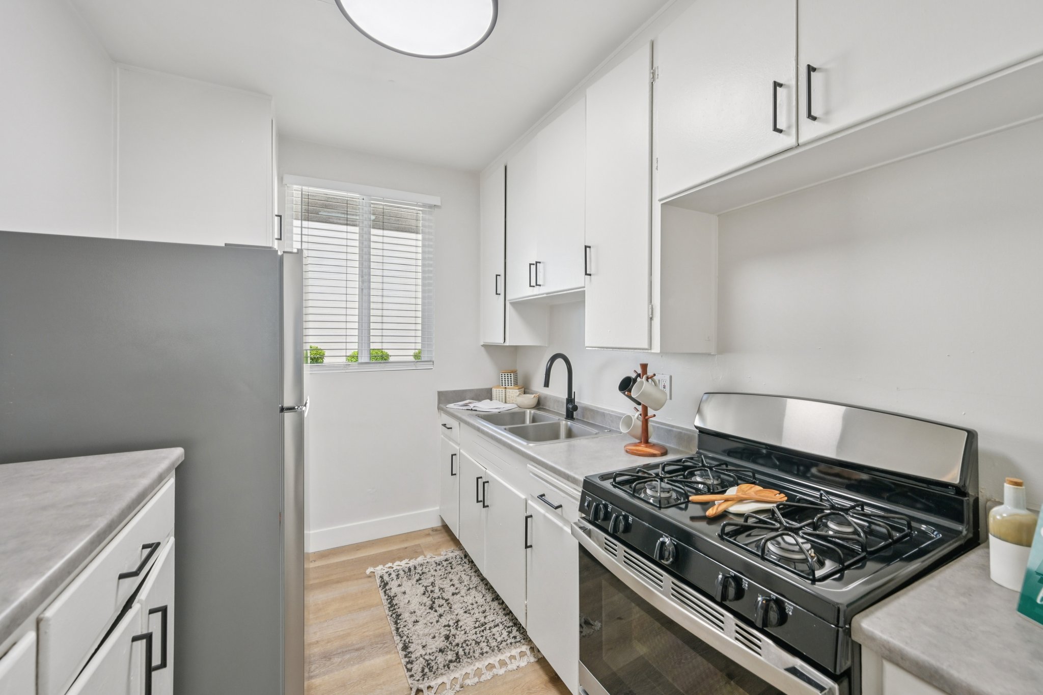 Modern kitchen with white cabinets, stainless steel appliances, and a gas stove. A refrigerator is against the left wall, with a sink next to it. Natural light comes in through a window, and there’s a small decorative rug on the wooden floor. The countertop is tidy, with minimal items displayed.