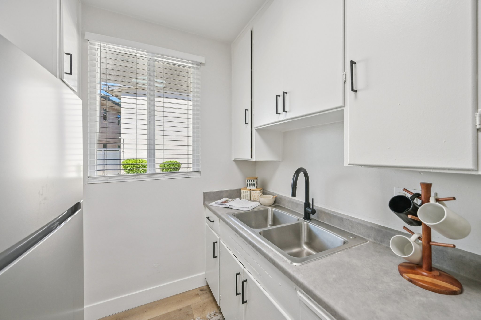 A bright, modern kitchen featuring white cabinets, a stainless steel sink, and a sleek black faucet. A window with blinds allows natural light to enter, and a mug tree with several cups is positioned on the countertop beside it. The flooring is light wood, contributing to the fresh aesthetic.