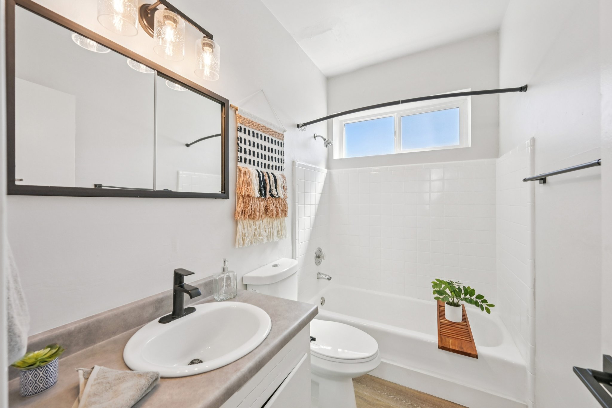 A bright and modern bathroom featuring a white bathtub with a shower, a large mirror above a sink with a black faucet, and decorative items including a plant and towel rack. The walls are painted white, and natural light enters through a window.