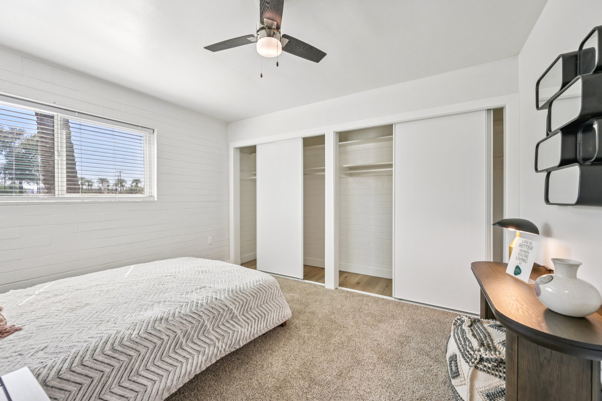 Bright and airy bedroom featuring a large window with blinds, a ceiling fan, and a neatly made bed with a textured blanket. The space includes a minimalist nightstand with a lamp and decorative items, as well as sliding closet doors on one side, enhancing the room's open and uncluttered feel.