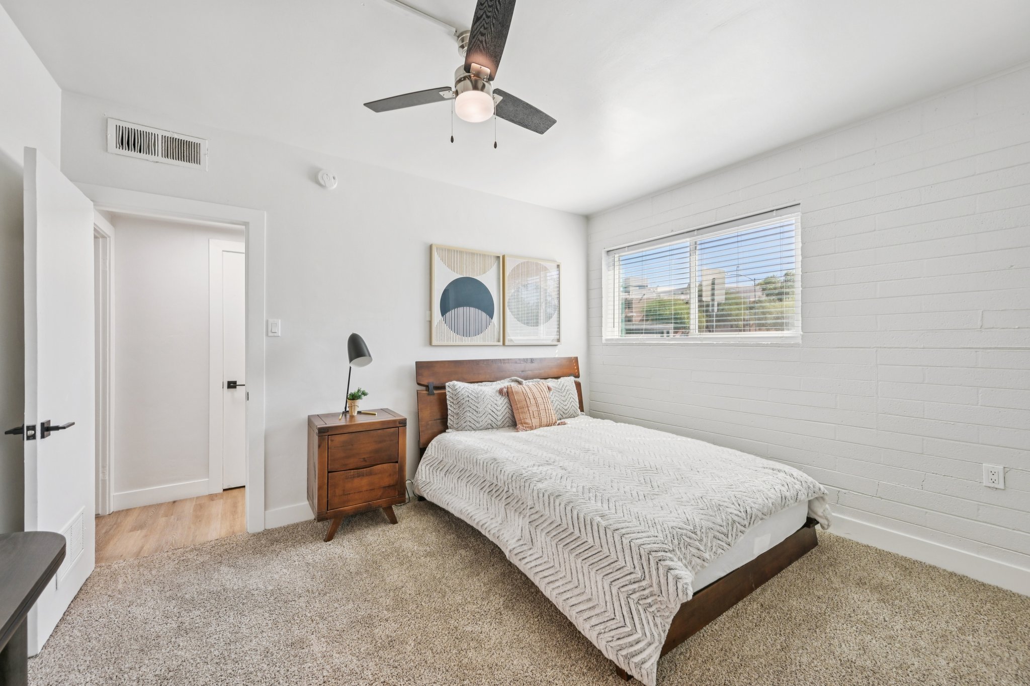 A spacious and modern bedroom with a queen-sized bed featuring a patterned white and gray blanket. There is a wooden nightstand with a lamp beside the bed, a window allowing natural light, and a decorative wall art piece above the headboard. The walls are painted white, and the floor is carpeted.