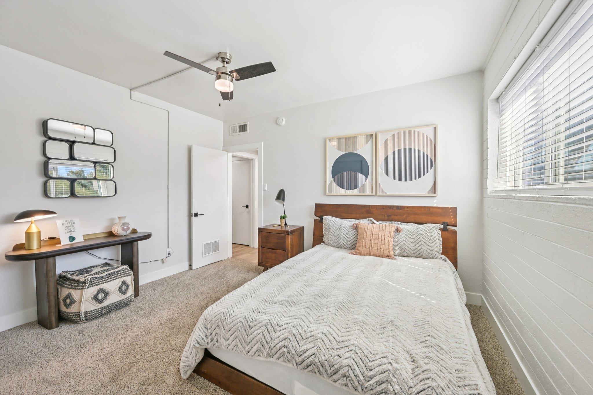 A cozy bedroom featuring a large bed with a textured blanket, a wooden nightstand, and a stylish lamp. The walls are light-colored, and large windows allow natural light to fill the space. Two framed abstract artworks are mounted above the bed, adding a modern touch. A small table and decorative basket complete the decor.
