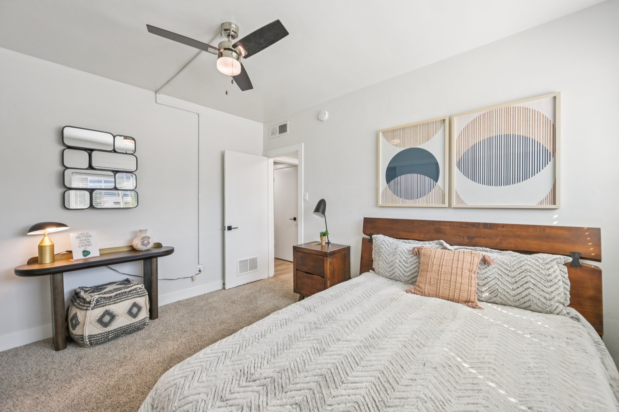 A cozy bedroom featuring a stylish bed with a patterned blanket, a nightstand with a lamp, a console table with decorative items, and two framed abstract art pieces on the wall. A ceiling fan adds functionality, and a light neutral color palette creates a serene atmosphere.