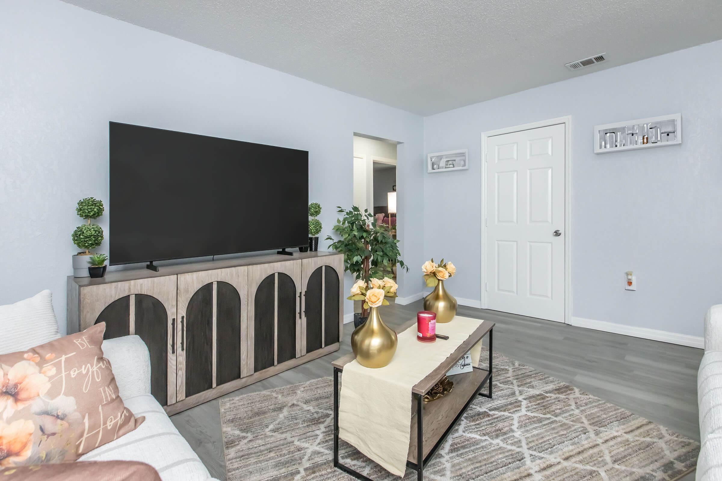 A modern living room featuring a large flat-screen TV mounted on a light blue wall. There is a wooden TV stand with decorative elements, including green plants and gold vases with yellow flowers. A coffee table with a neutral table runner sits in the center, complemented by a red candle. Light-colored sofas complete the inviting space.
