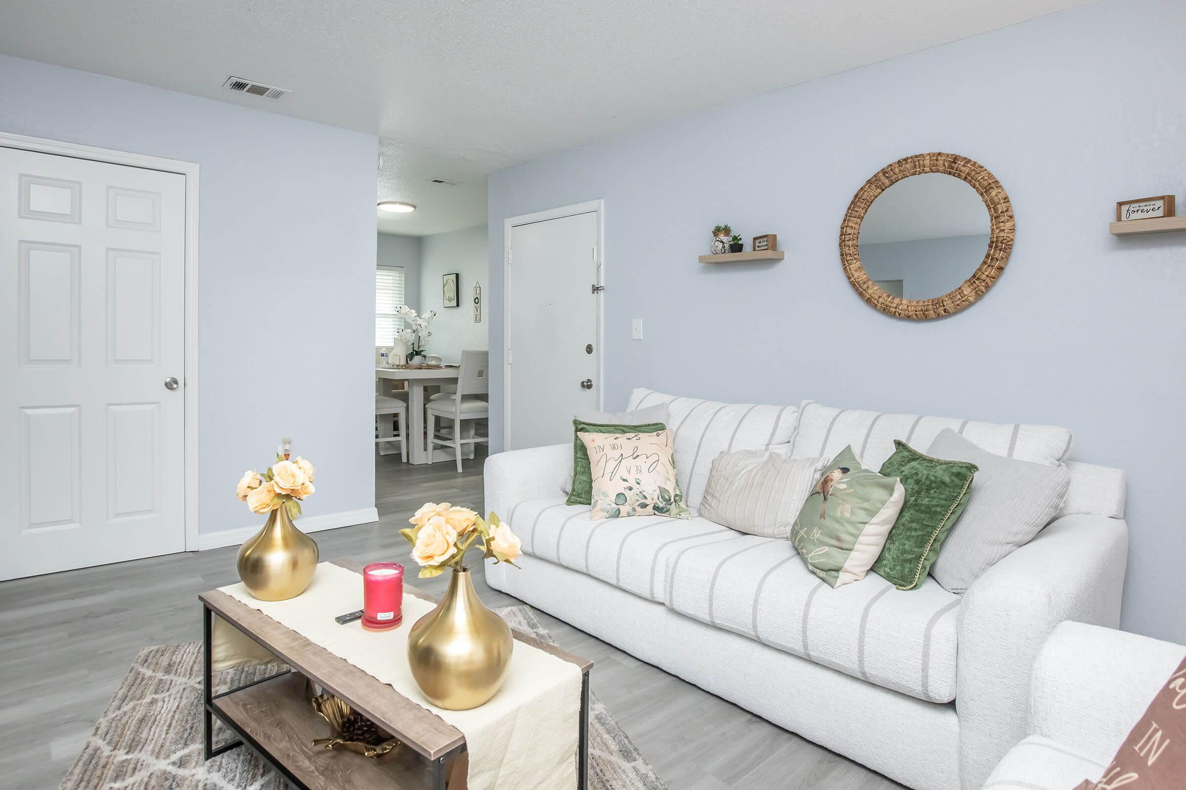 A cozy living room featuring a light gray sofa adorned with decorative pillows, two gold-colored vases with yellow flowers on a coffee table, and a circular mirror on the wall. A white door leads to another room, and a dining area with a light wooden table and chairs can be seen in the background.