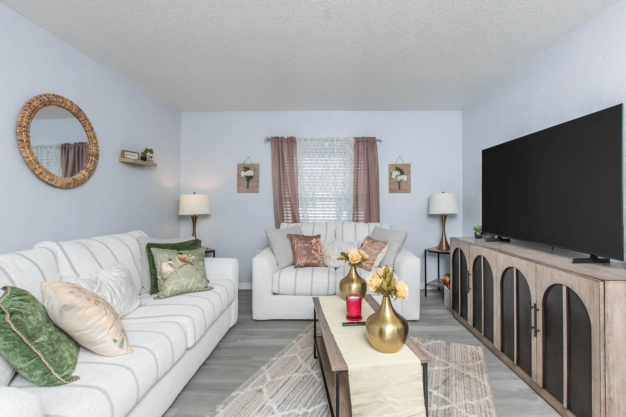 Cozy living room featuring a light-colored sofa and armchair with decorative pillows. A coffee table with vases of flowers sits in the center. The room has a large TV on a wood cabinet, two lamps, a round mirror, and light curtains, creating a warm and inviting atmosphere.