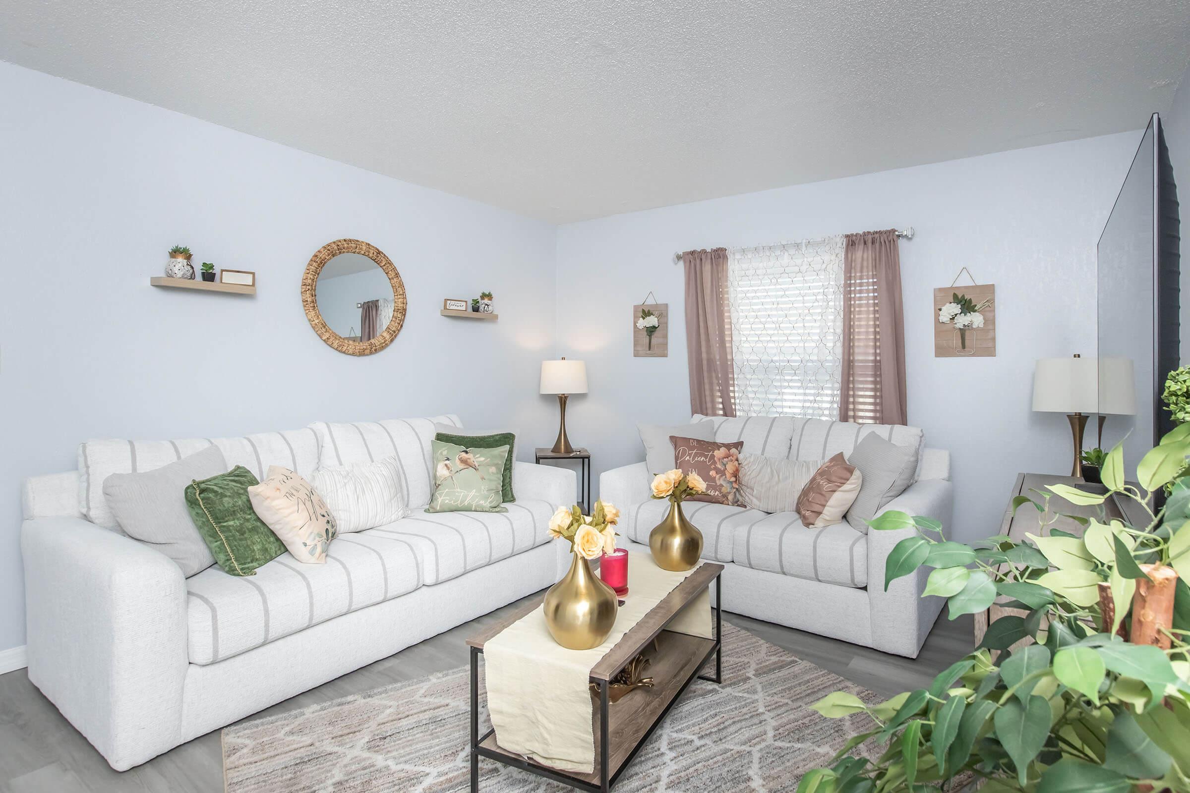 A bright and cozy living room featuring two white sofas adorned with decorative pillows. A wooden coffee table holds vases with flowers, and a soft area rug is beneath. Wall decorations include framed floral art and shelves with small plants. Natural light filters through a window covered with sheer curtains.
