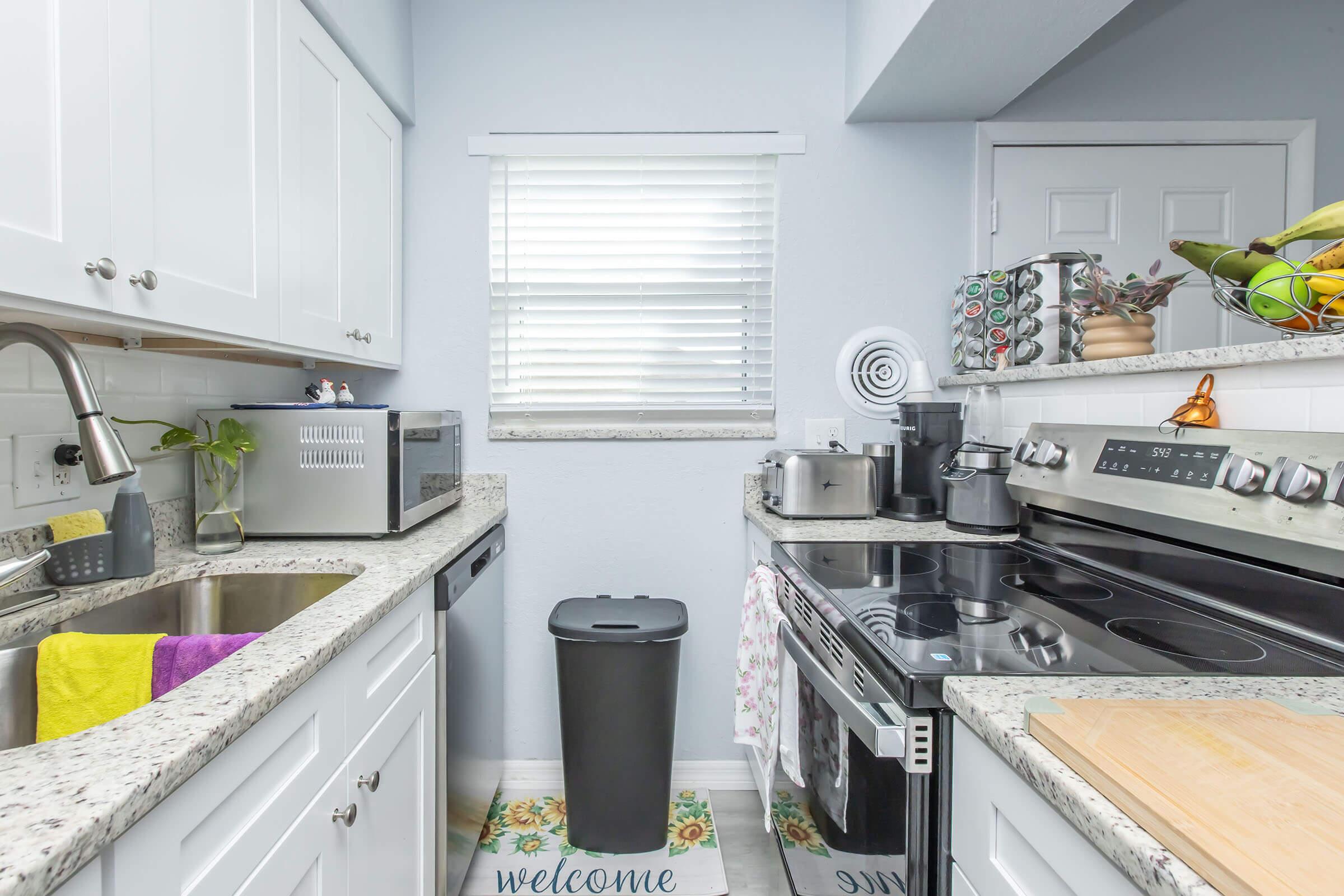 A modern kitchen featuring white cabinetry, a stainless steel stove, and a sink with a granite countertop. There is a trash can, kitchen utensils, and a decorative welcome mat on the floor. A window with blinds allows natural light to fill the space, enhancing the clean and tidy appearance.