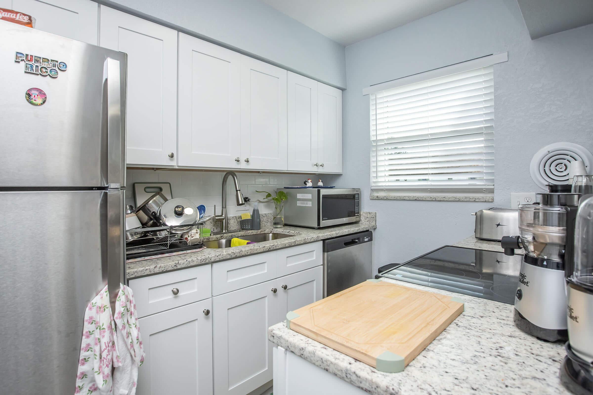 A modern kitchen featuring white cabinetry, a granite countertop, a stainless steel refrigerator, a microwave, and a stove. There are dishes in a drying rack near the sink. A wooden cutting board rests on the counter, and light streams in through a window with white blinds.