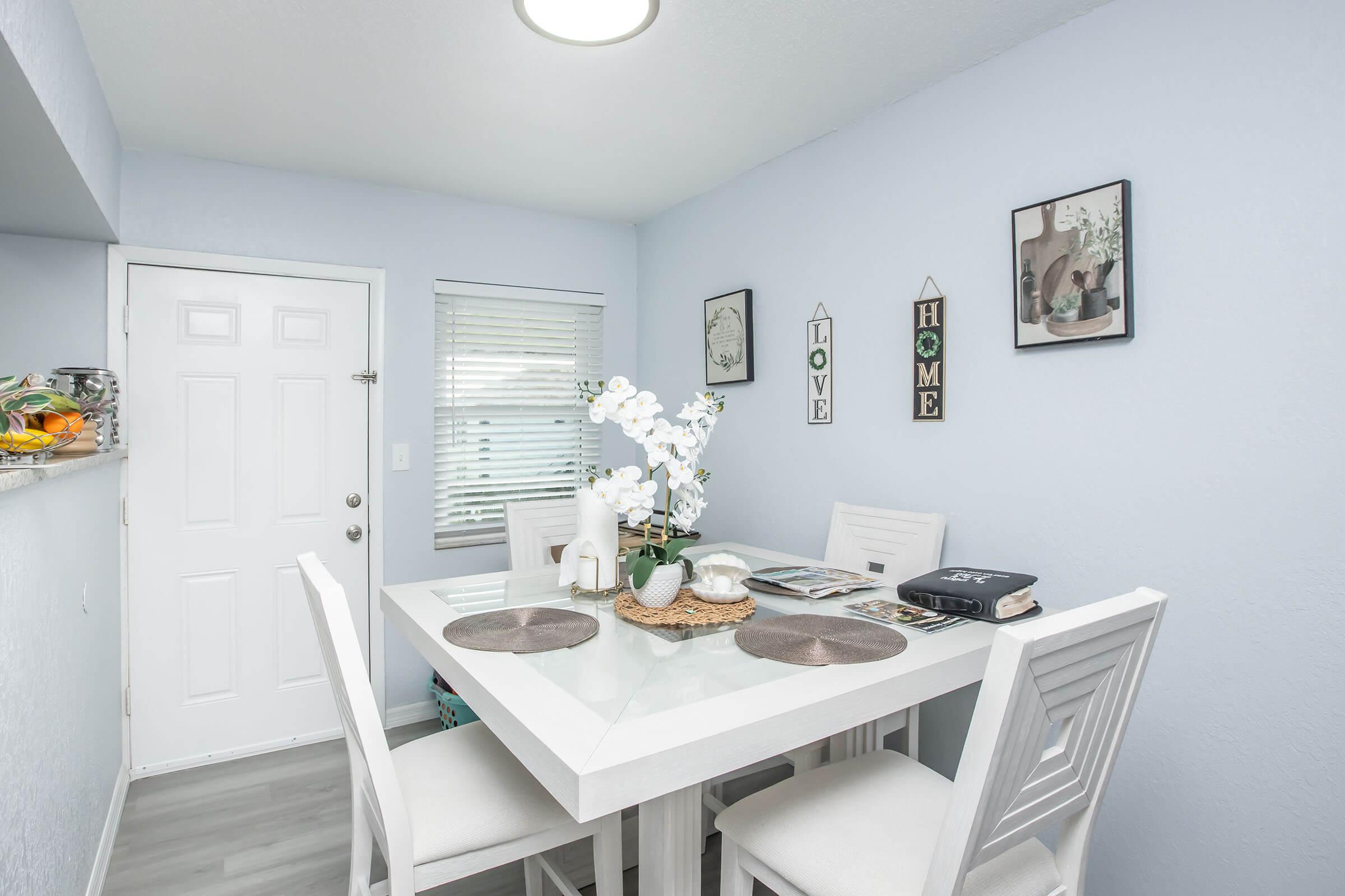 A bright dining area featuring a white table with four matching chairs, set with placemats and a magazine. A window with blinds allows natural light, while a door leads outside. The walls are painted light blue, adorned with framed artwork and a vase of white orchids on the table, creating a cozy and inviting atmosphere.