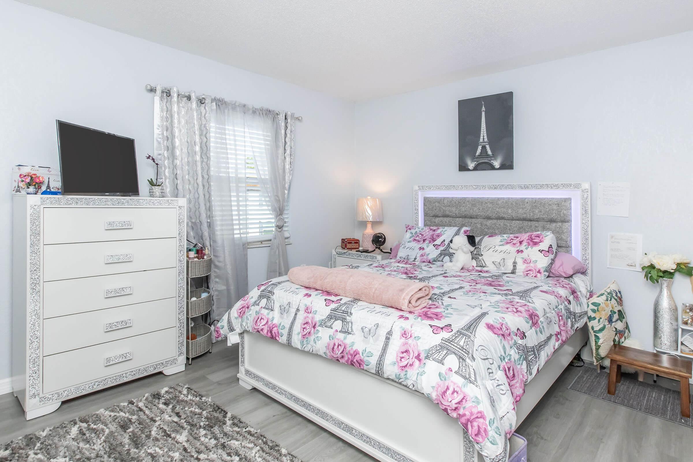 A cozy bedroom featuring a floral-patterned bedspread on a bed with a pink blanket. There's a white dresser with ornate details, a television on a stand, and light-colored curtains. A small vase of flowers and a picture of the Eiffel Tower add charm to the decor. The room has a light and airy feel.