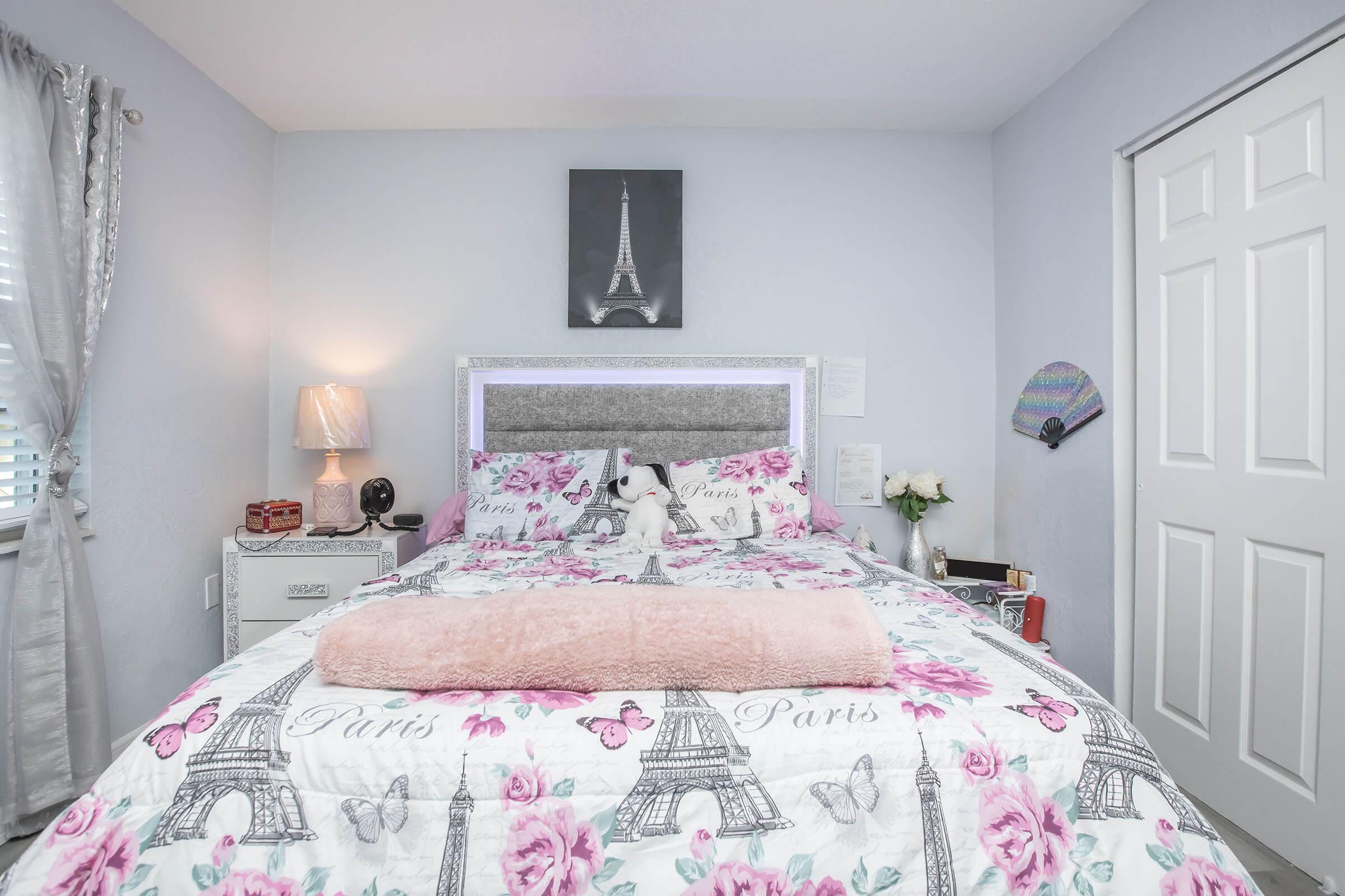 A cozy bedroom featuring a bed with a floral Paris-themed quilt, pink throw blanket, and a stuffed animal. There’s a nightstand with a lamp and a flower vase. A wall art of the Eiffel Tower is above the bed, and a closed wardrobe is visible in the background with light-colored walls and curtains.