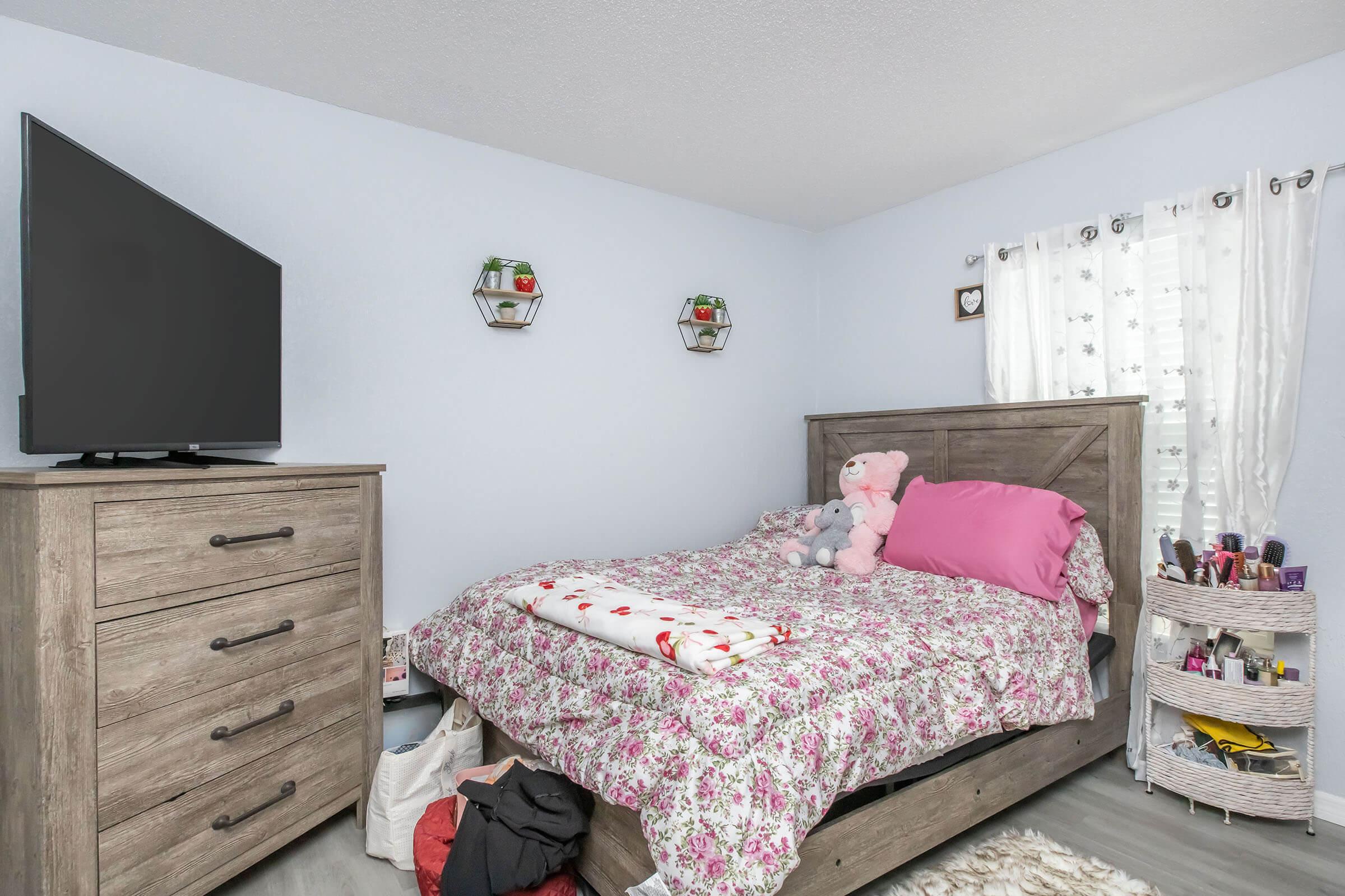 A cozy bedroom featuring a wooden bed with floral bedding, a pink pillow, and stuffed animals. There's a dresser with a television on top, alongside wall-mounted shelves with decorative items. A window with sheer curtains lets in light, and a small makeup organizer sits beside the bed.
