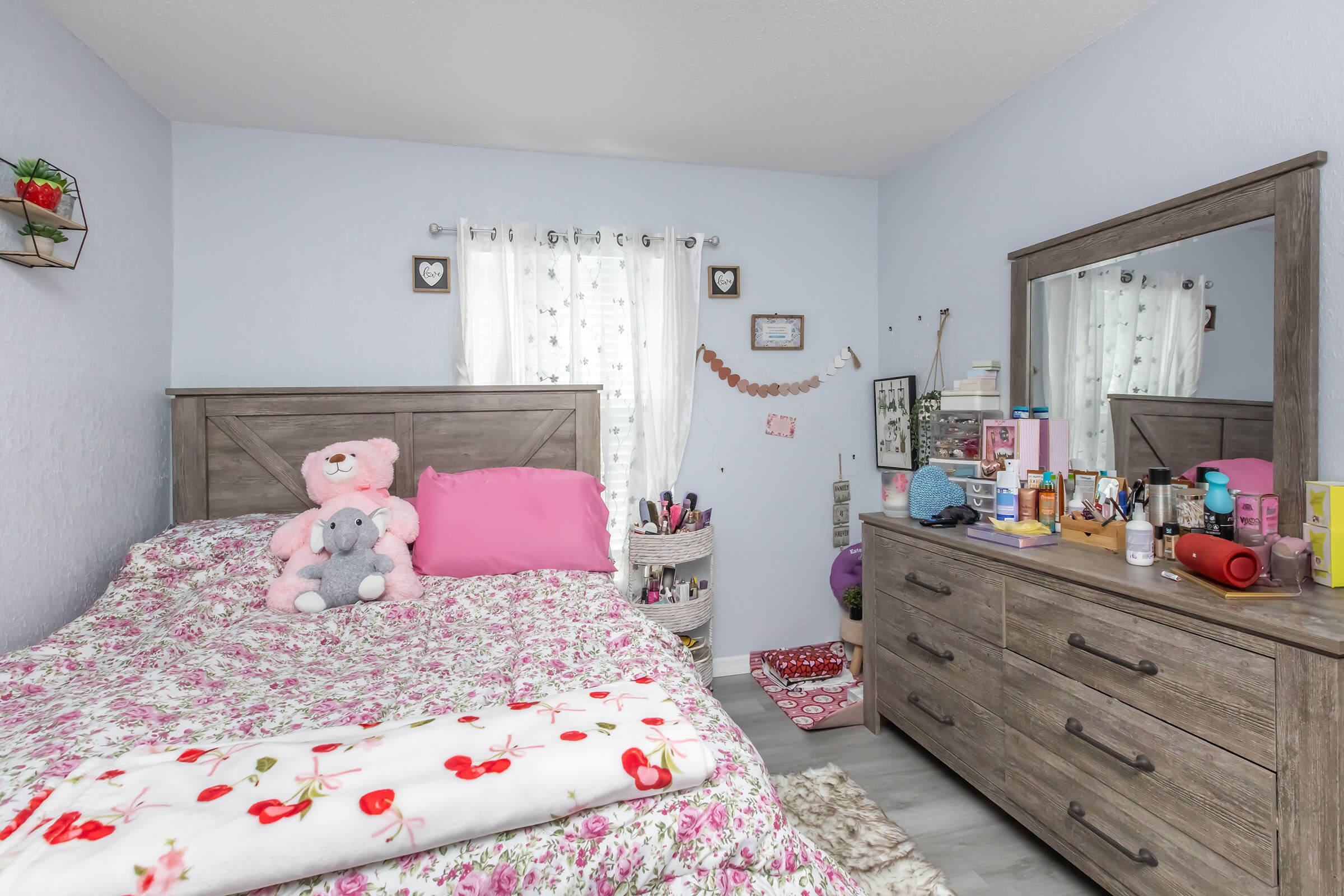 A cozy bedroom featuring a bed with floral bedding and a pink teddy bear, a wooden dresser adorned with beauty products, and a window with light curtains. Decorative items and personal touches create a warm atmosphere. The room is well-organized and inviting.