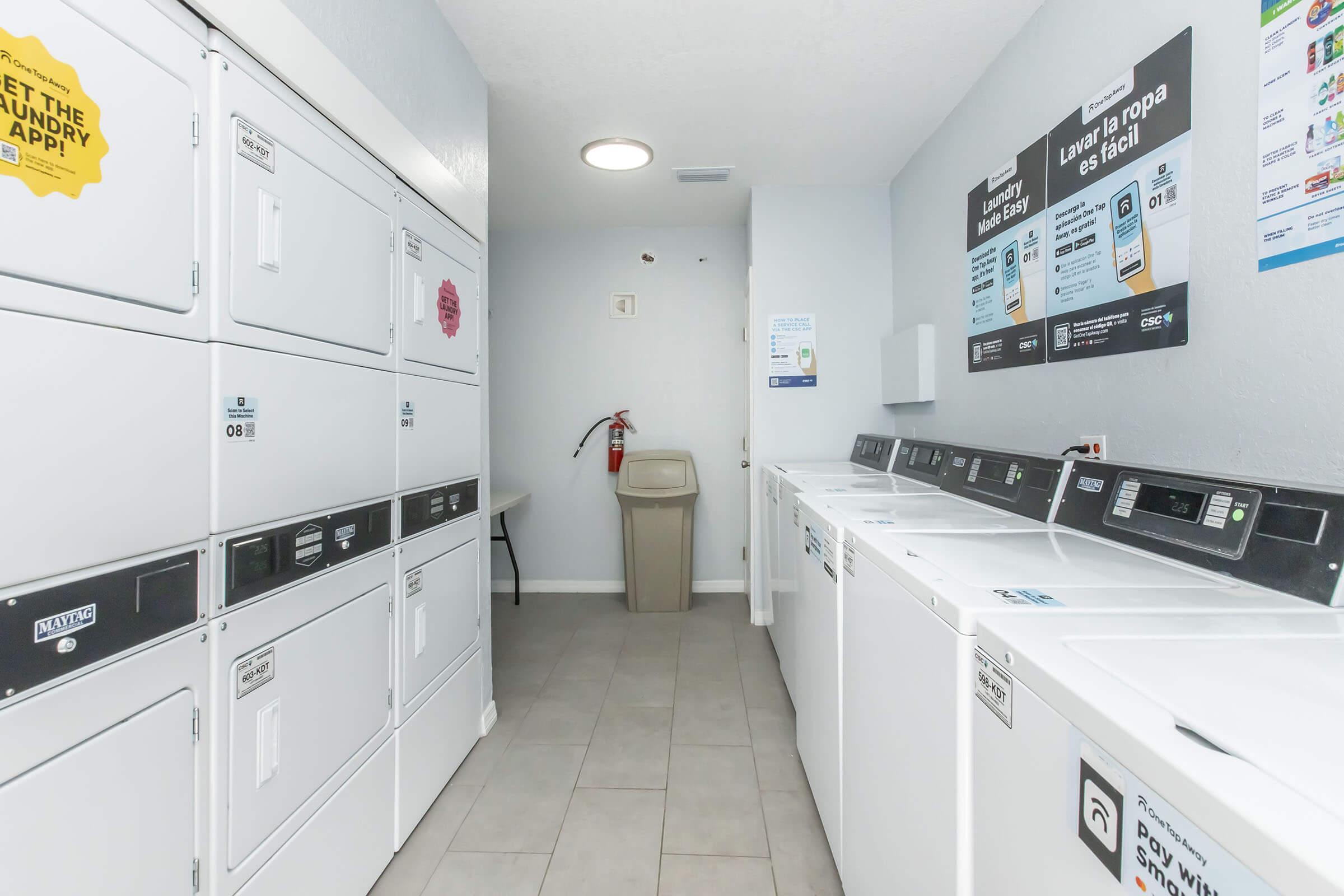 A clean and well-lit laundry room featuring several stacked washing machines and dryers against the wall. There is a trash bin and a fire extinguisher visible. Informational signs are posted on the wall, providing instructions and services related to laundry. The floor is tiled and the space looks organized.