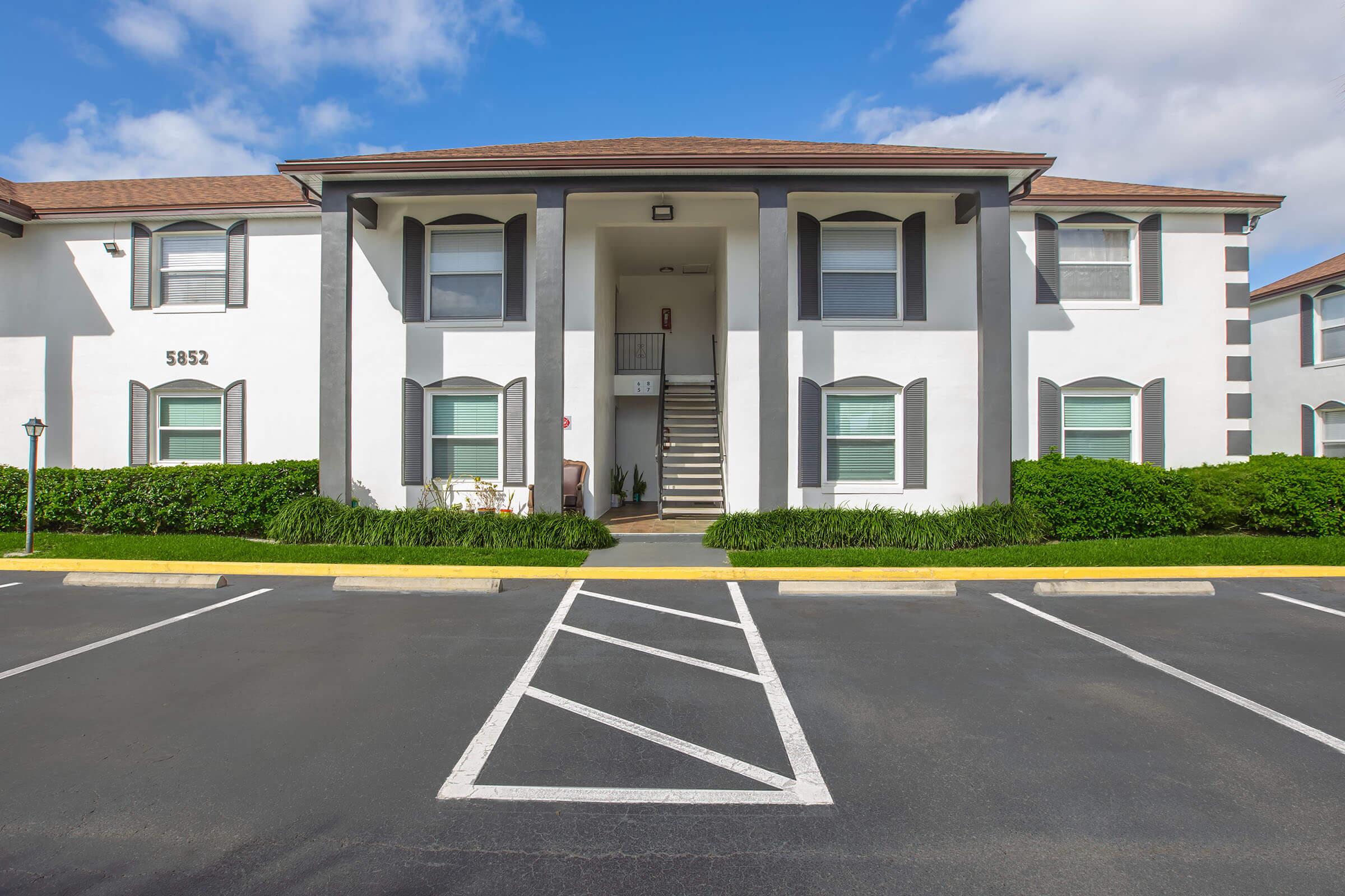 A two-story apartment building with a white exterior and dark gray accents. There are stairs leading to the entrance, flanked by large windows. The surrounding landscape features green shrubbery and a clear blue sky. Parking spaces are visible in front of the building, with a few marked lines.