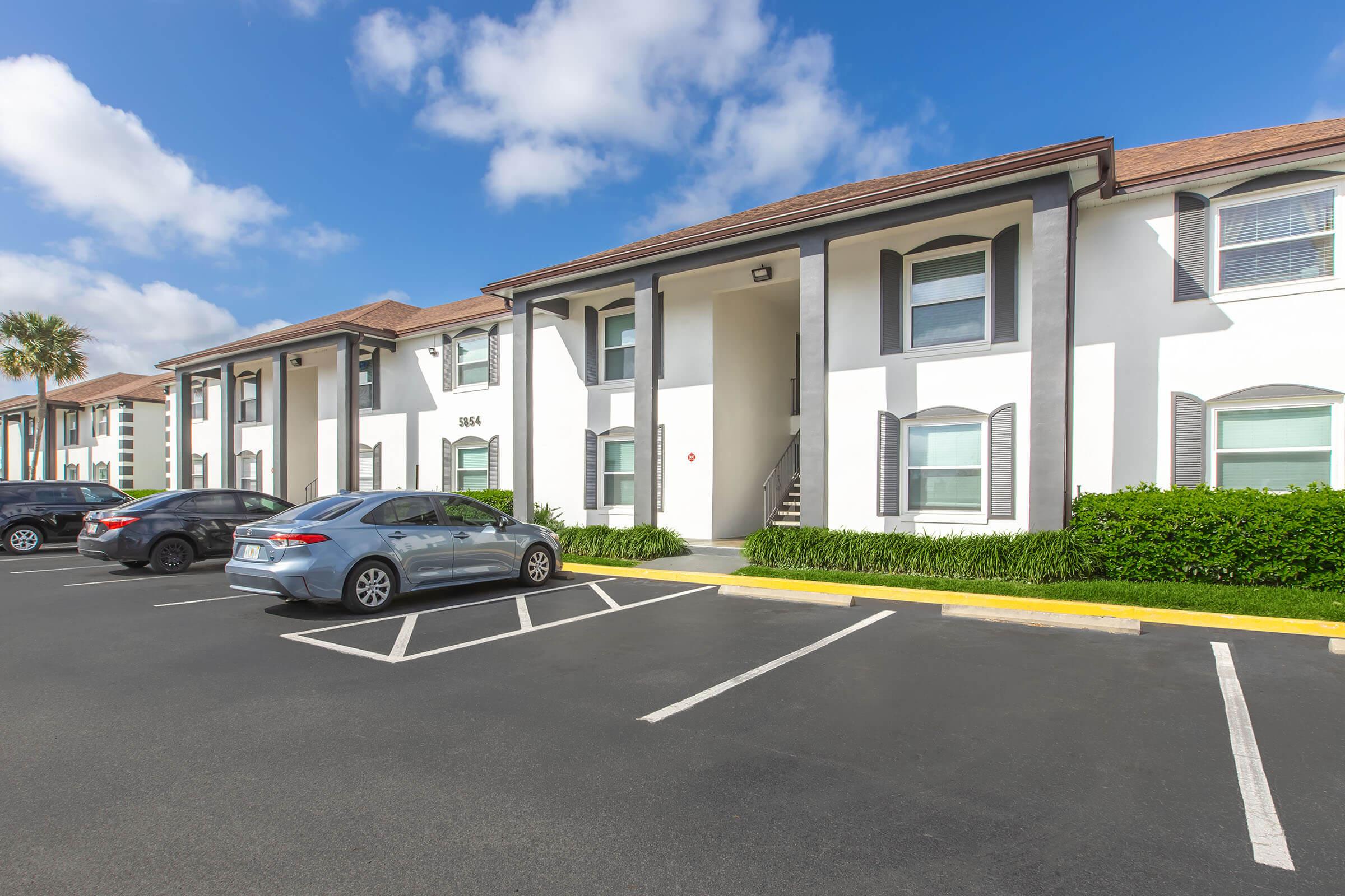 A modern apartment complex featuring multiple units with white walls and gray accents. The image shows parking spaces, a well-maintained landscape with green shrubs, and a clear blue sky with scattered clouds. A few parked cars are visible along the front of the building.