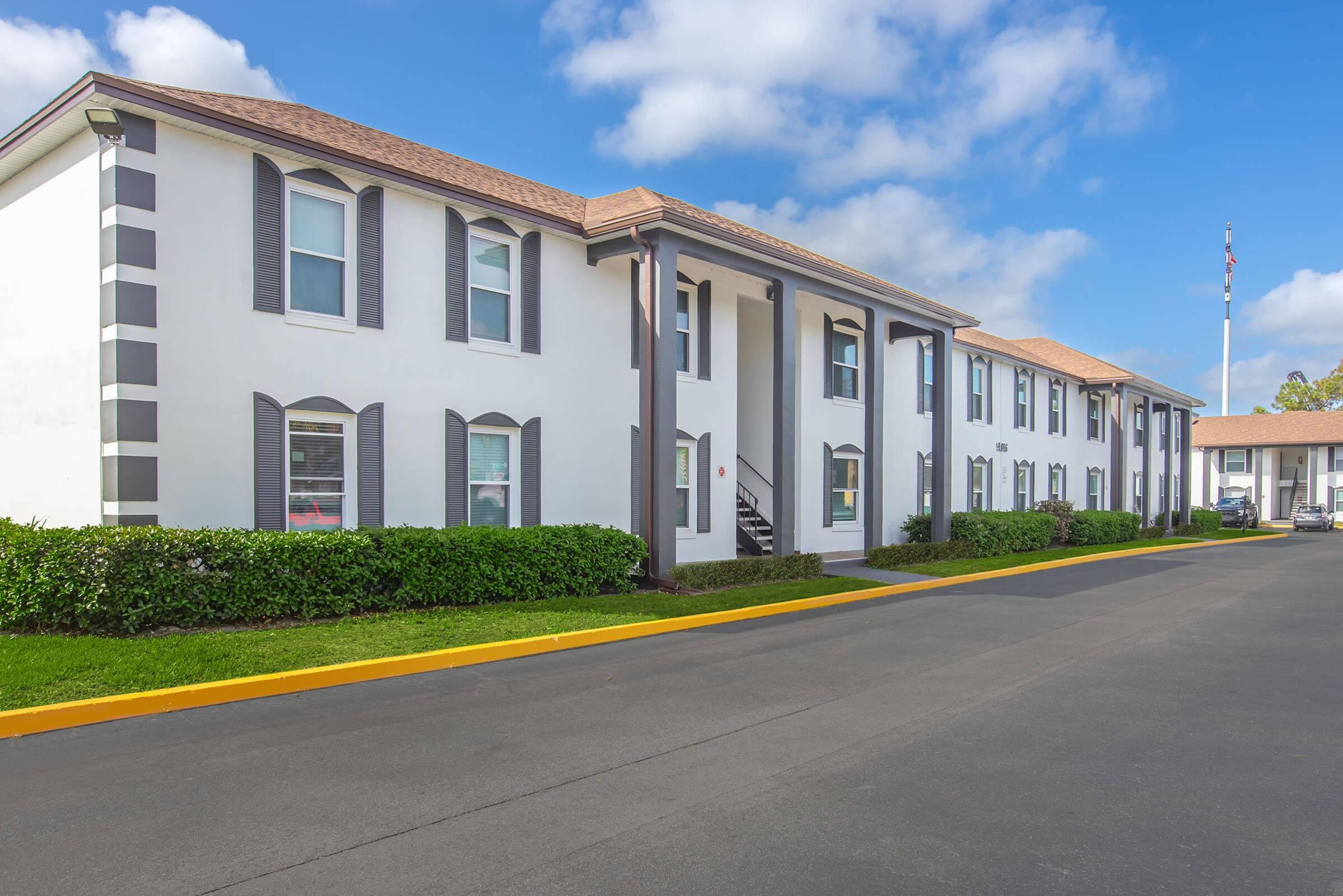 A two-story building with a white exterior and dark gray accents, featuring large windows and a staircase. The foreground includes a neatly landscaped area with shrubs, and the road is paved. Blue skies with scattered clouds are visible above the structure.