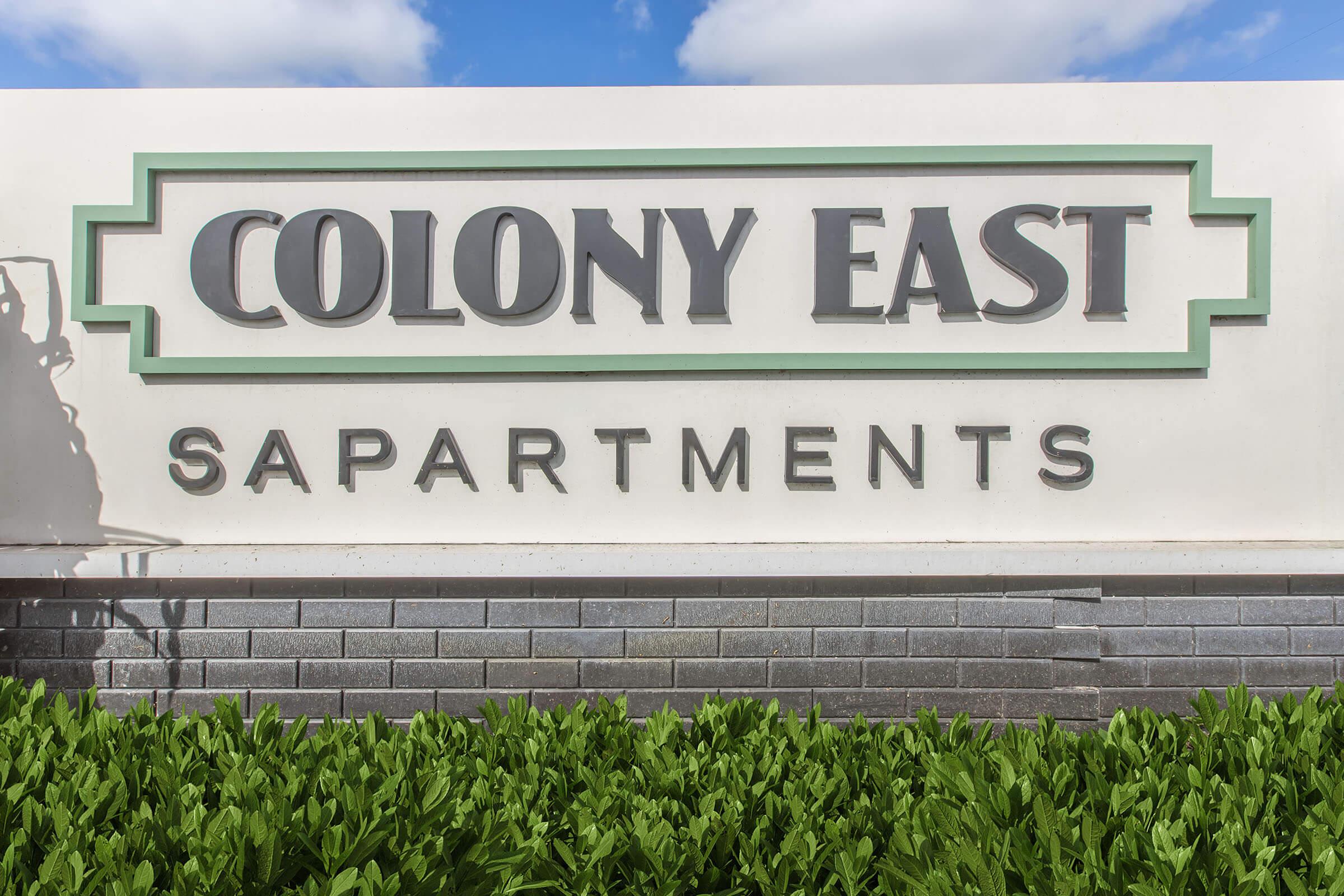 Signage for Colony East Apartments, featuring the name "COLONY EAST" prominently displayed in large letters with decorative elements, situated above the word "APARTMENTS." The sign is positioned against a backdrop of green foliage and a clear blue sky.