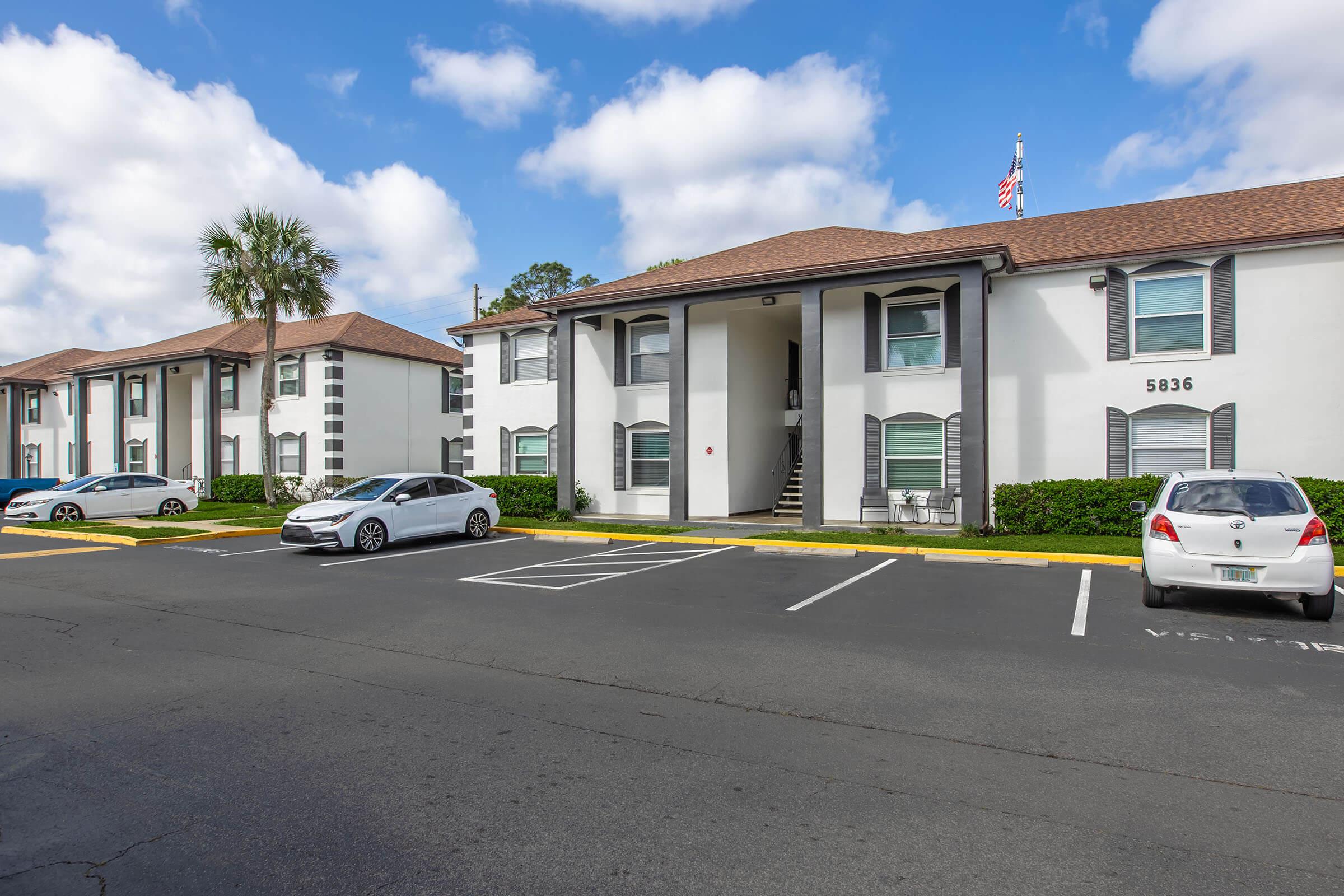A residential apartment complex featuring two buildings with white exteriors and brown roofs. There are several parked cars in the foreground and a staircase leading to an entrance. A flag is visible on one building, with partly cloudy blue skies above.