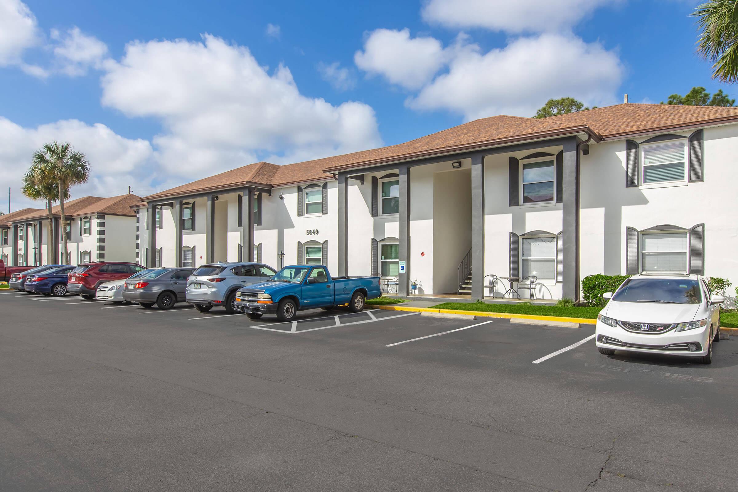 A white two-story apartment building with brown shingles, featuring multiple parking spaces filled with various cars. A clear blue sky with scattered clouds is overhead. The scene includes palm trees and well-maintained landscaping.