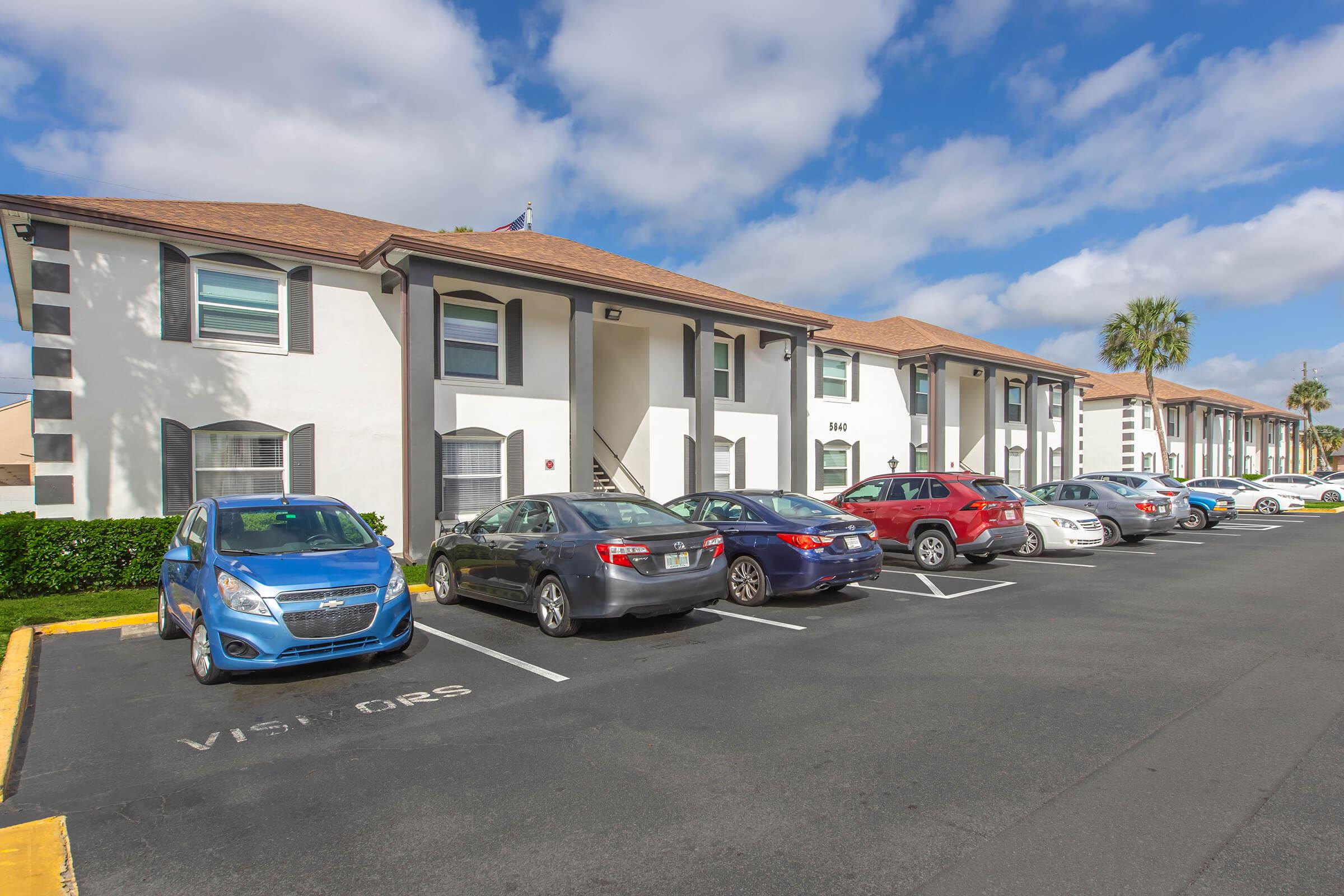 A well-maintained apartment complex featuring two-story white buildings with brown roofs. Multiple parked cars, including a blue hatchback and a red sedan, line the parking lot. Lush green landscaping is visible in front of the buildings under a partly cloudy sky.