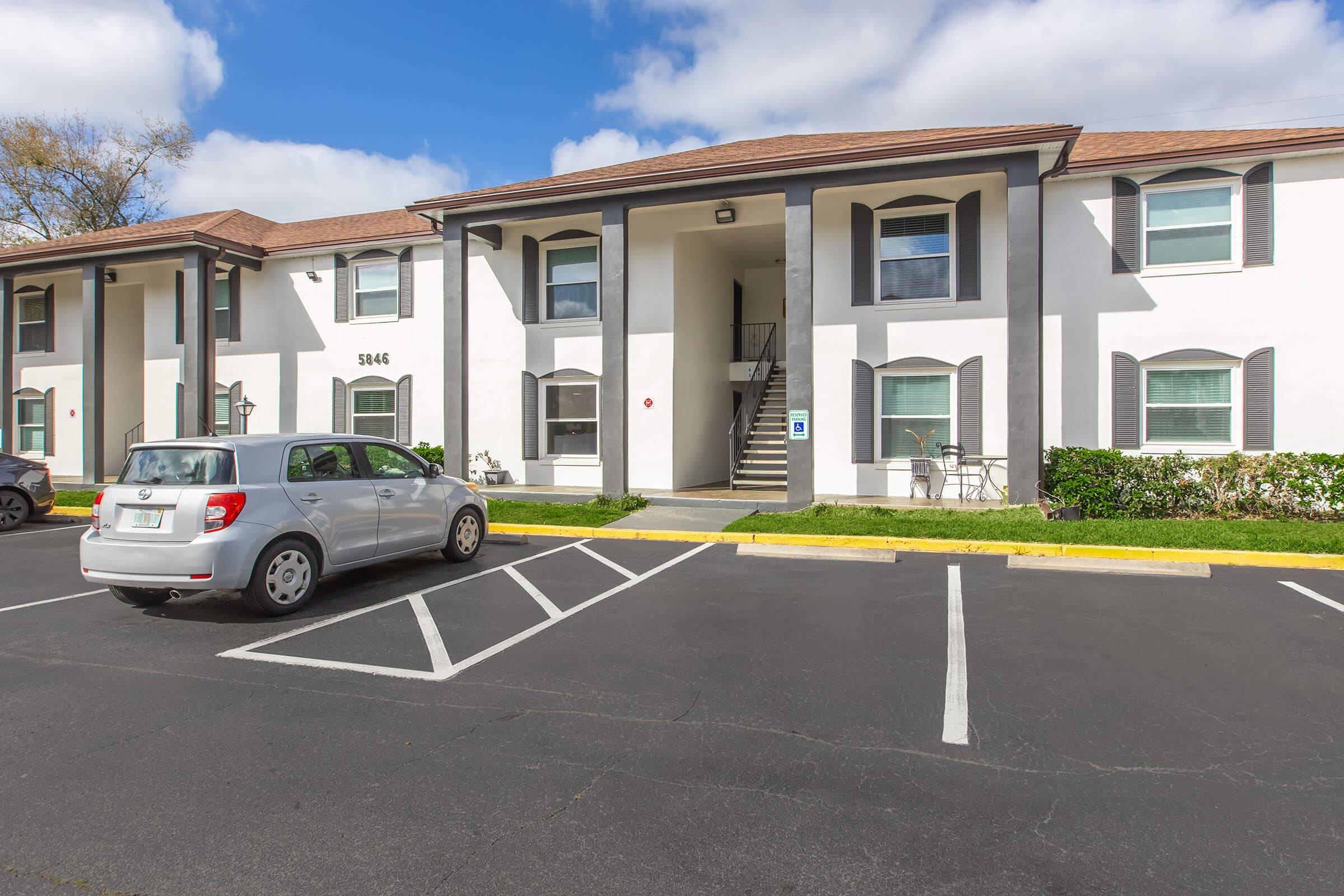 Exterior view of a two-story residential building with a white facade and brown roof. A silver car is parked in the foreground, with several parking spaces visible. The entrance features a staircase, and there are green shrubs and a small seating area outside the building. Bright blue sky with some clouds in the background.