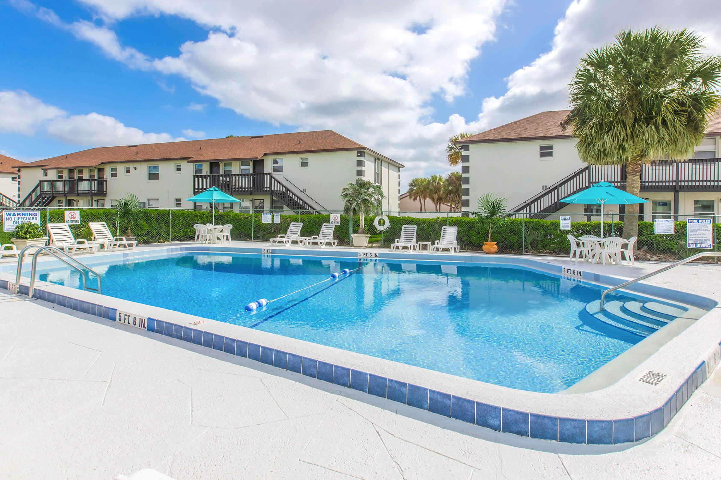 A clear swimming pool surrounded by lounge chairs and umbrellas, located in a residential area. Two-story apartment buildings are visible in the background, with green landscaping and palm trees. The sky is partly cloudy, creating a bright and inviting atmosphere.