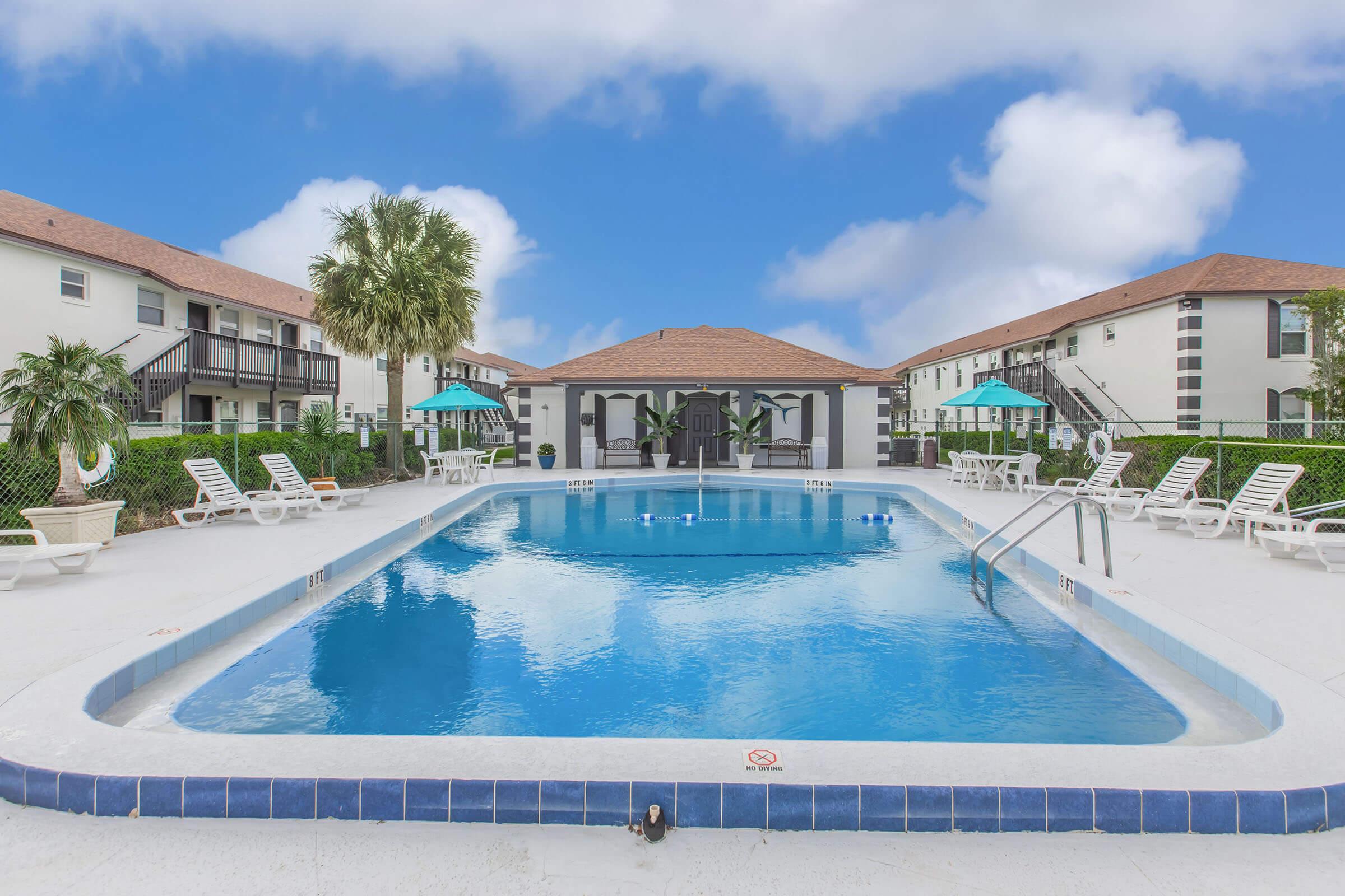 A crystal-clear swimming pool surrounded by lounge chairs and palm trees, with two buildings featuring green umbrellas in the background. The sky is partly cloudy, creating a serene and inviting atmosphere.