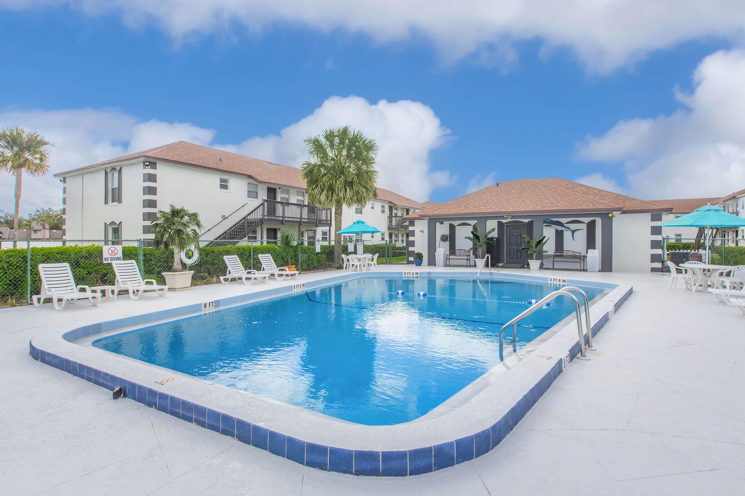 A clear blue swimming pool surrounded by lounge chairs and tropical greenery. In the background, a two-story building with a red tiled roof and umbrella-covered seating areas. The sky is bright and cloudy, creating a serene and inviting atmosphere.