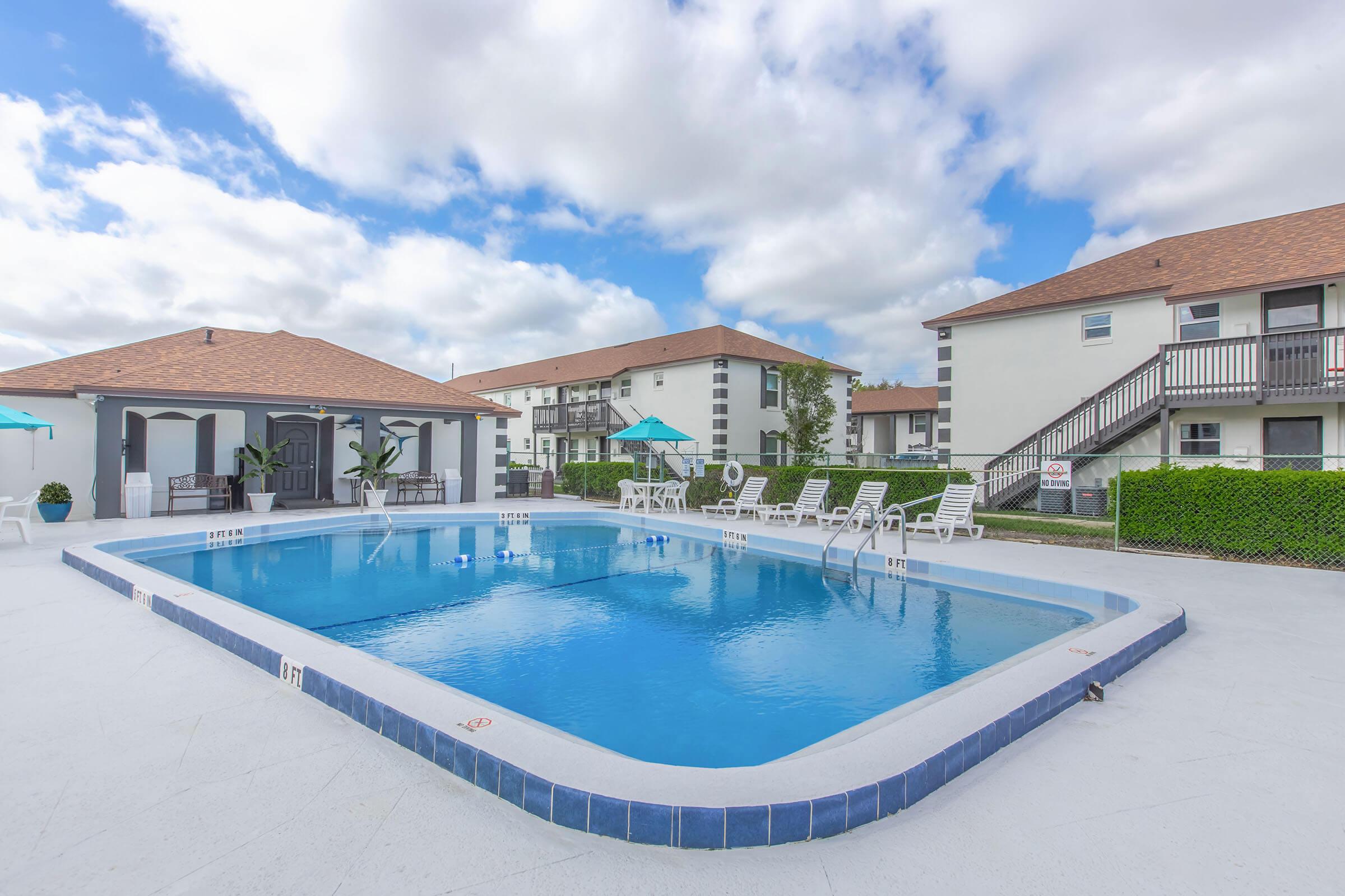 A large swimming pool surrounded by lounge chairs, with umbrellas for shade. In the background, two buildings with a light-colored facade and brown roofs can be seen under a partly cloudy sky. The pool area has a clean, inviting look, perfect for relaxation and recreation.