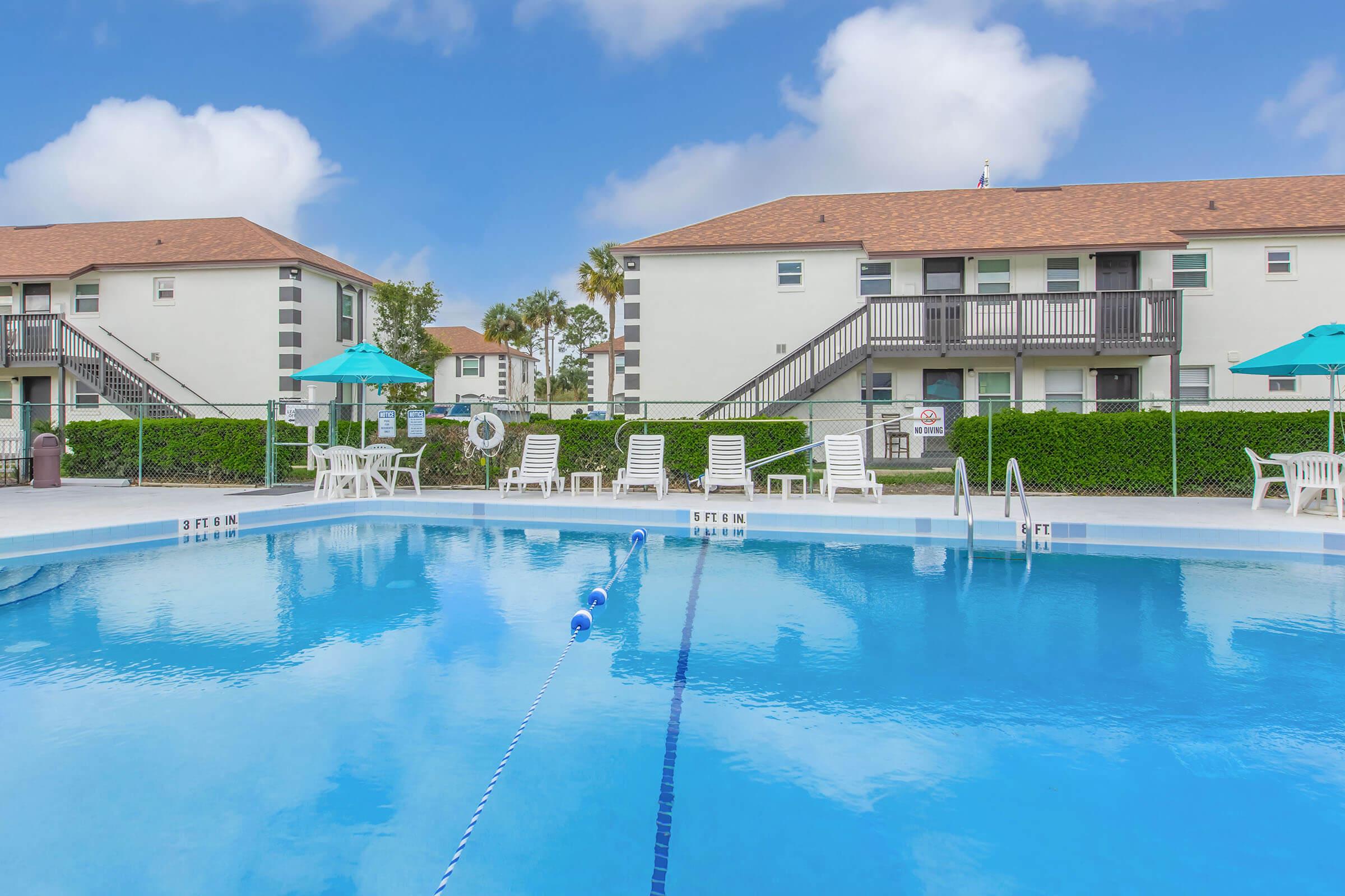 A clear blue swimming pool surrounded by lounge chairs and umbrellas, with two apartment buildings in the background. The sky is bright with fluffy white clouds, and the pool area is well-maintained, featuring diving lanes and a landscaped border.