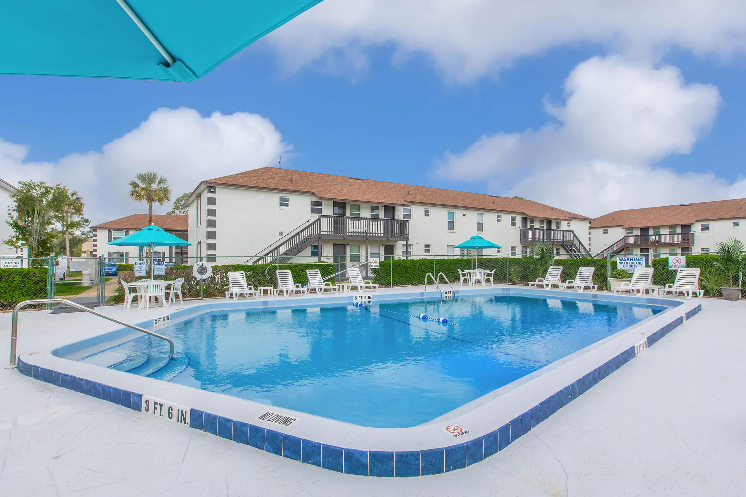 A clear blue swimming pool surrounded by white lounge chairs and umbrellas, set against a backdrop of two-story residential buildings. The area is landscaped with green bushes and palm trees, under a bright blue sky with scattered clouds. The pool depth is marked as 3 to 6 feet.