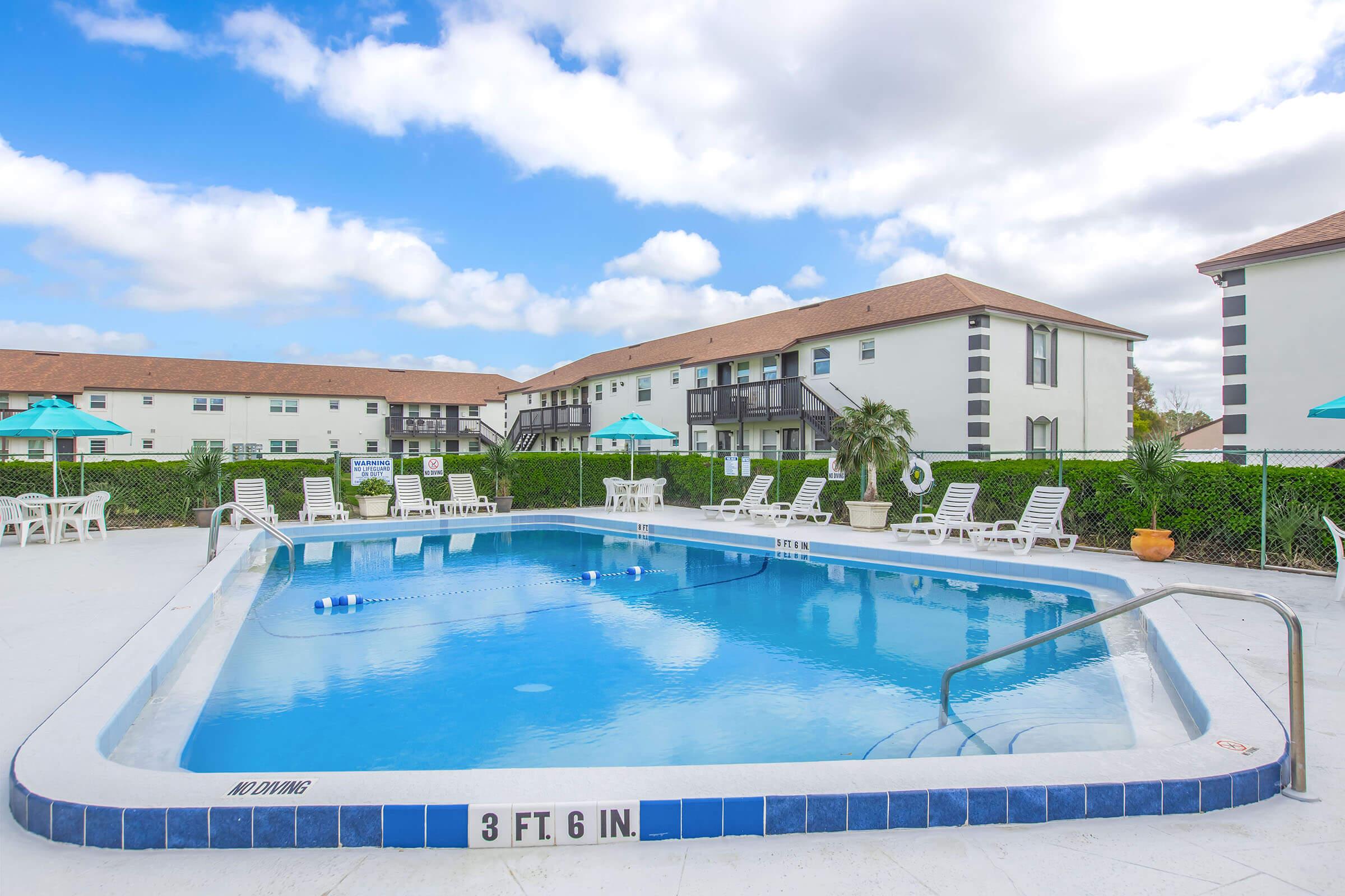 A clear blue swimming pool surrounded by white lounge chairs and umbrellas. In the background, there are two-story apartment buildings with a light color scheme. The sky is partly cloudy, creating a bright and inviting atmosphere. A depth marker shows the pool is 3 feet 6 inches deep.