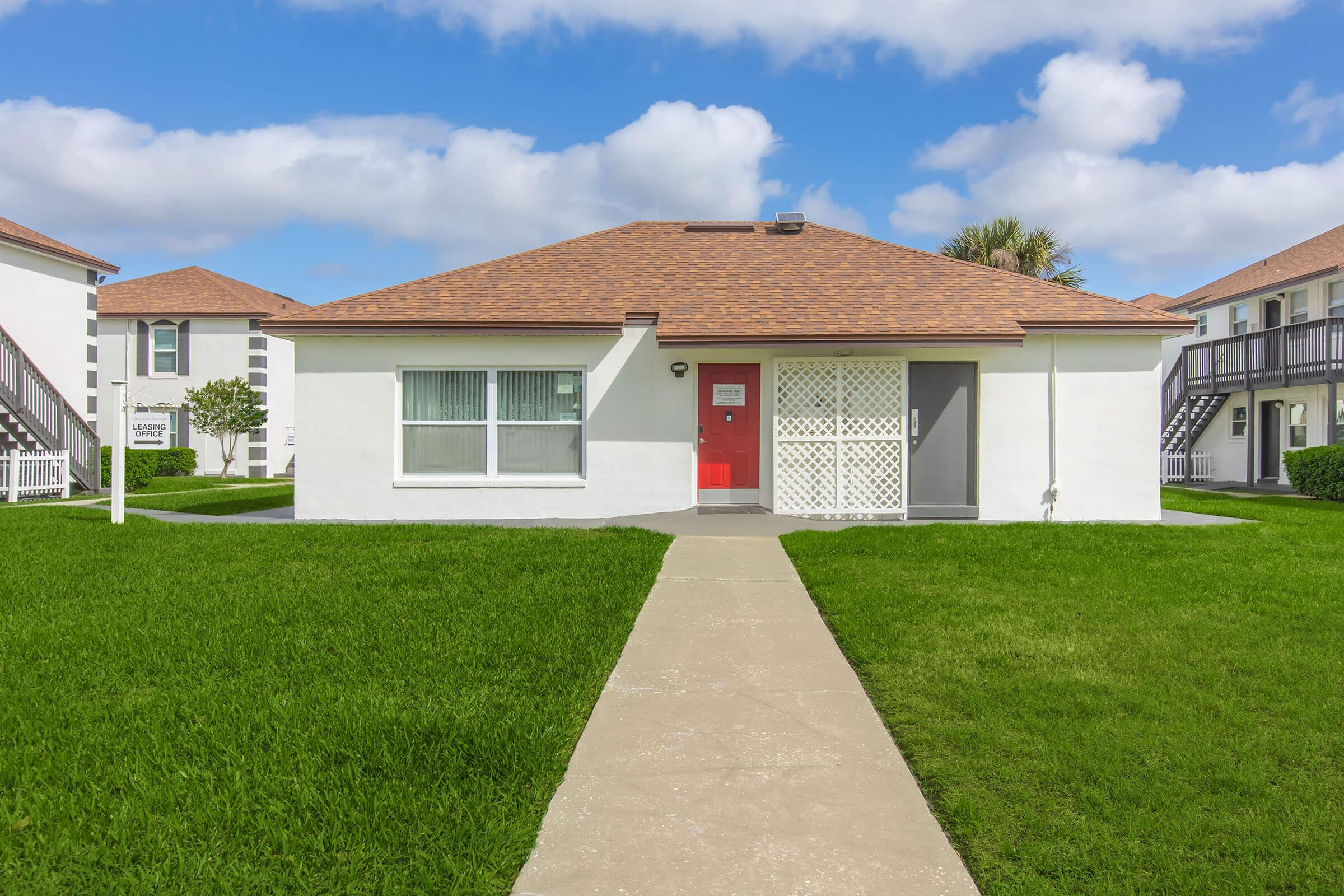 A single-story white house with a red door, featuring a latticework screen on one side. The house is surrounded by lush green grass and has a clear blue sky with fluffy white clouds in the background. There are two multi-unit buildings in the background with balconies, creating a pleasant residential setting.