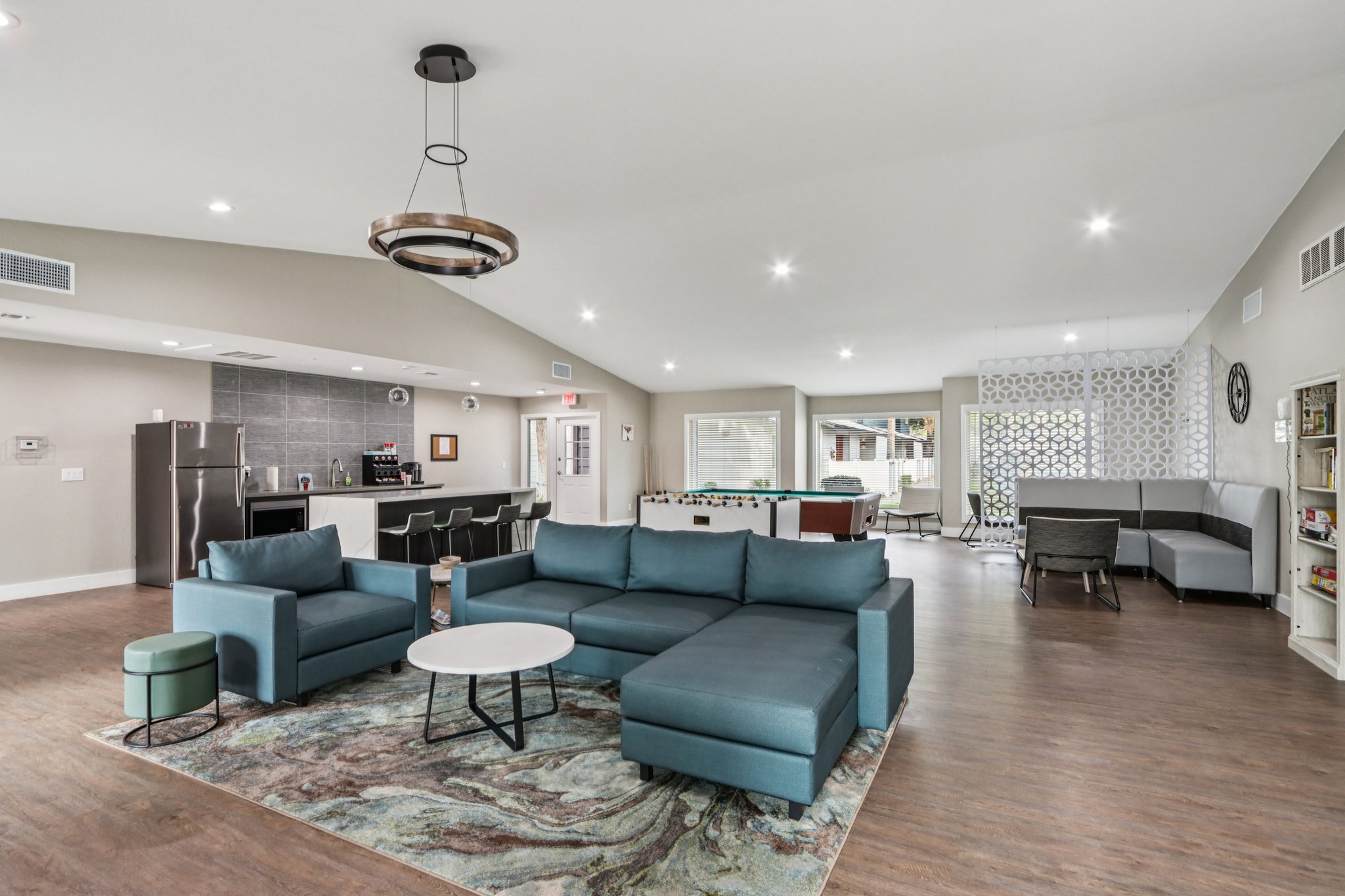 Spacious modern living area featuring a large sectional sofa, round coffee table, and a rug with abstract patterns. The background includes a kitchen area with stainless steel appliances and a bar counter. Bright natural lighting enhances the open atmosphere, with decorative elements and seating areas throughout.