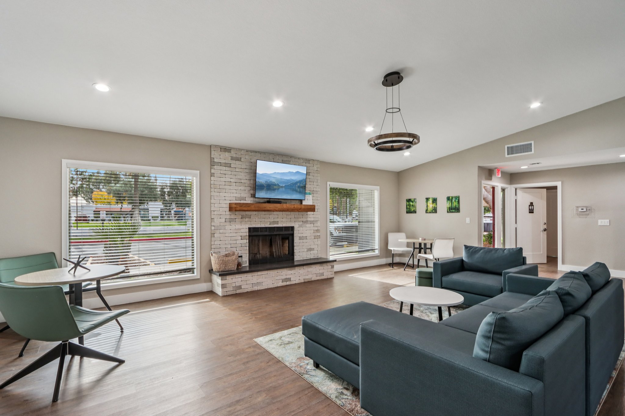 Modern living room with a comfortable blue sofa, two small round tables, and a cozy fireplace featuring a television above it. Large windows allow natural light to enter, showcasing a view of the outdoors. The walls are light-colored, with decorative artwork and plants enhancing the welcoming atmosphere.