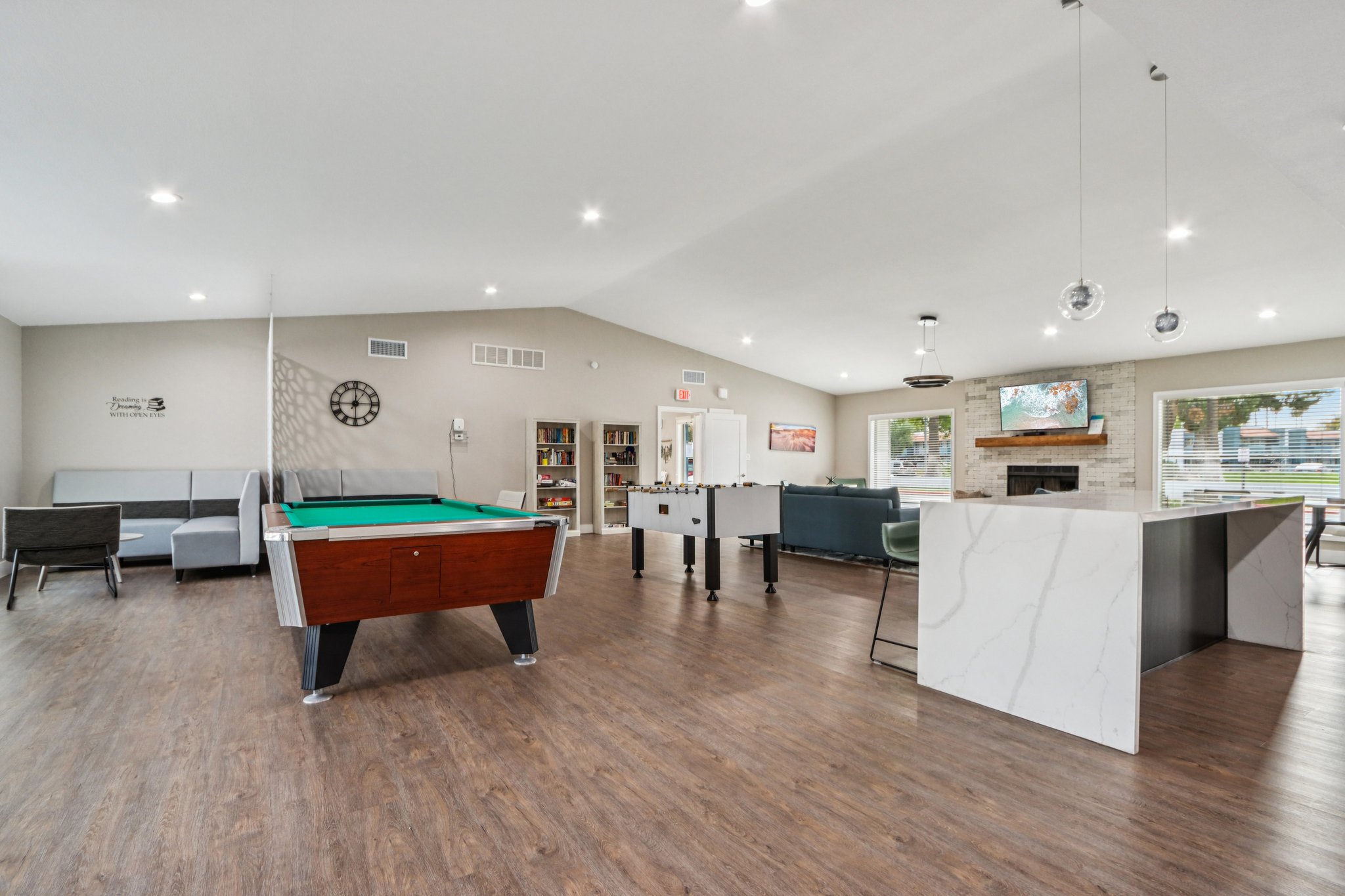 A modern communal lounge area featuring a billiard table, foosball table, and comfortable seating. Large windows offer natural light, while a stone fireplace adds warmth. Bookshelves and a clean, organized aesthetic enhance the inviting atmosphere. Ideal for socializing and relaxation.