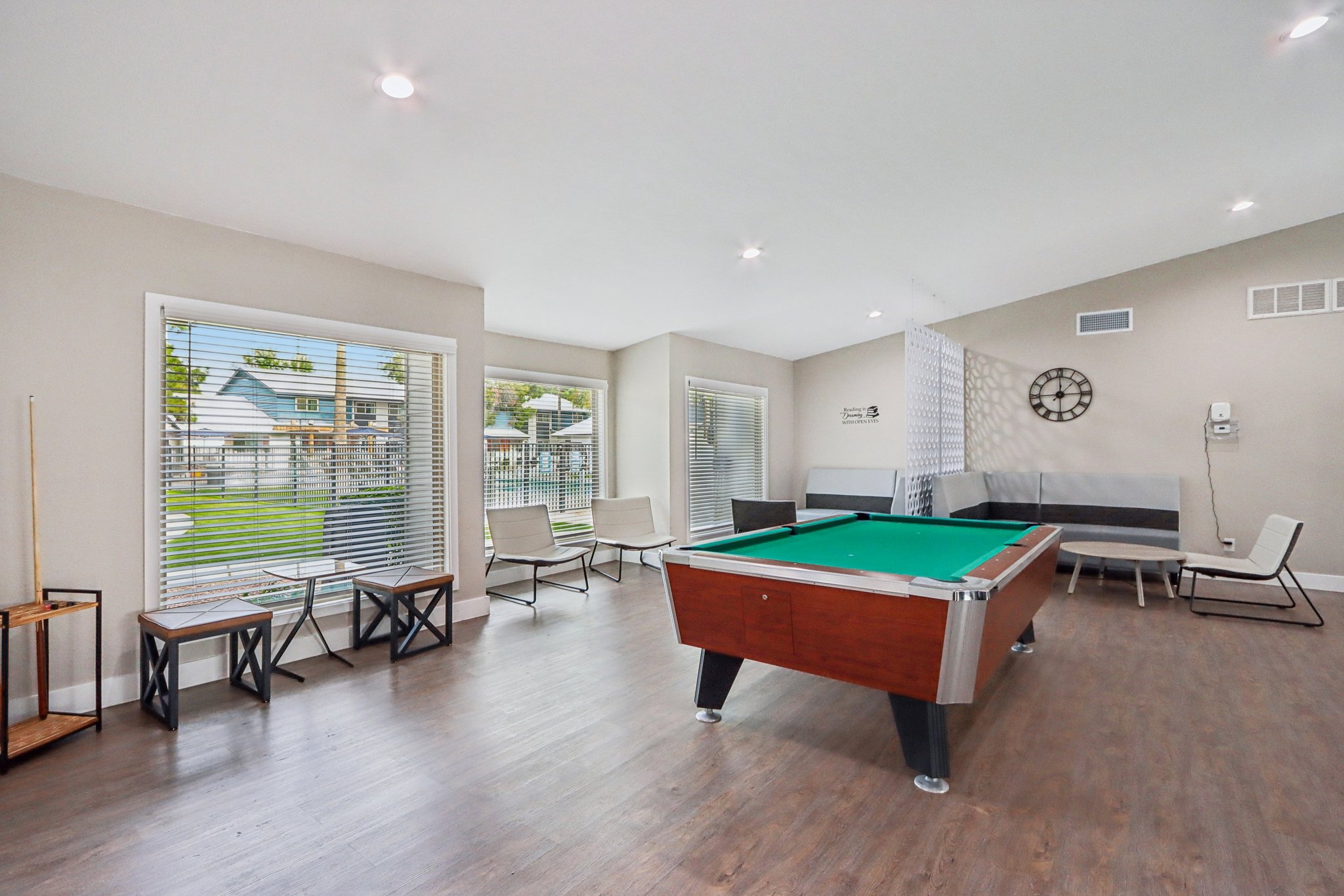 A bright, modern game room featuring a green billiards table at the center. Surrounding the table are stylish seating arrangements with a light-colored sofa and small tables. Large windows provide natural light and views of an outdoor space. The decor is clean and contemporary with neutral tones.