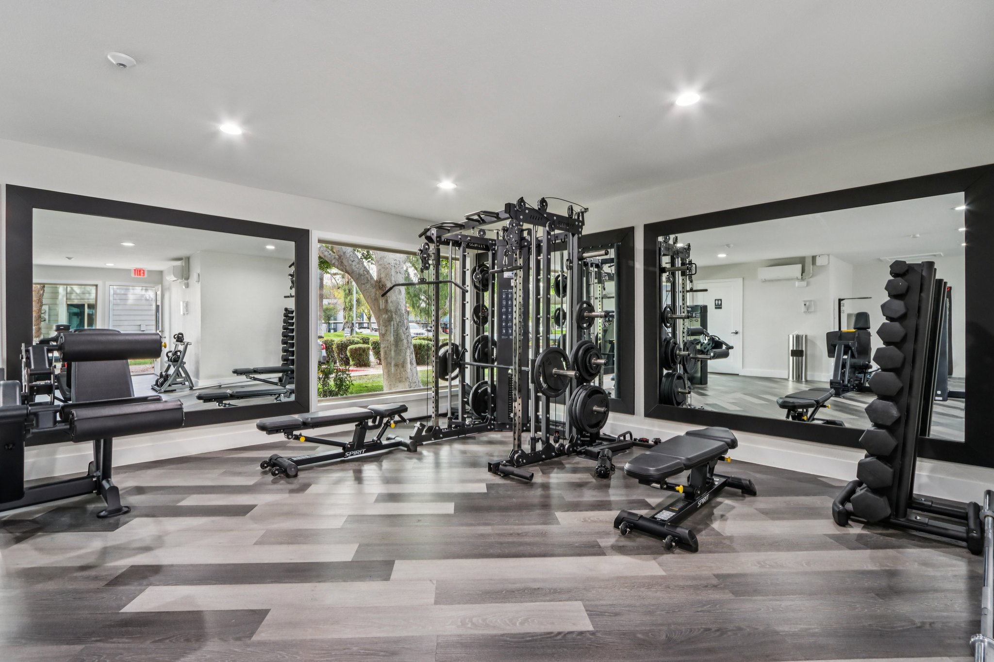 A modern gym interior featuring various fitness equipment, including weight benches, a multi-station gym, and free weights. Large mirrors reflect the space, enhancing the bright and open atmosphere. Natural light shines through windows, contributing to an inviting workout environment.