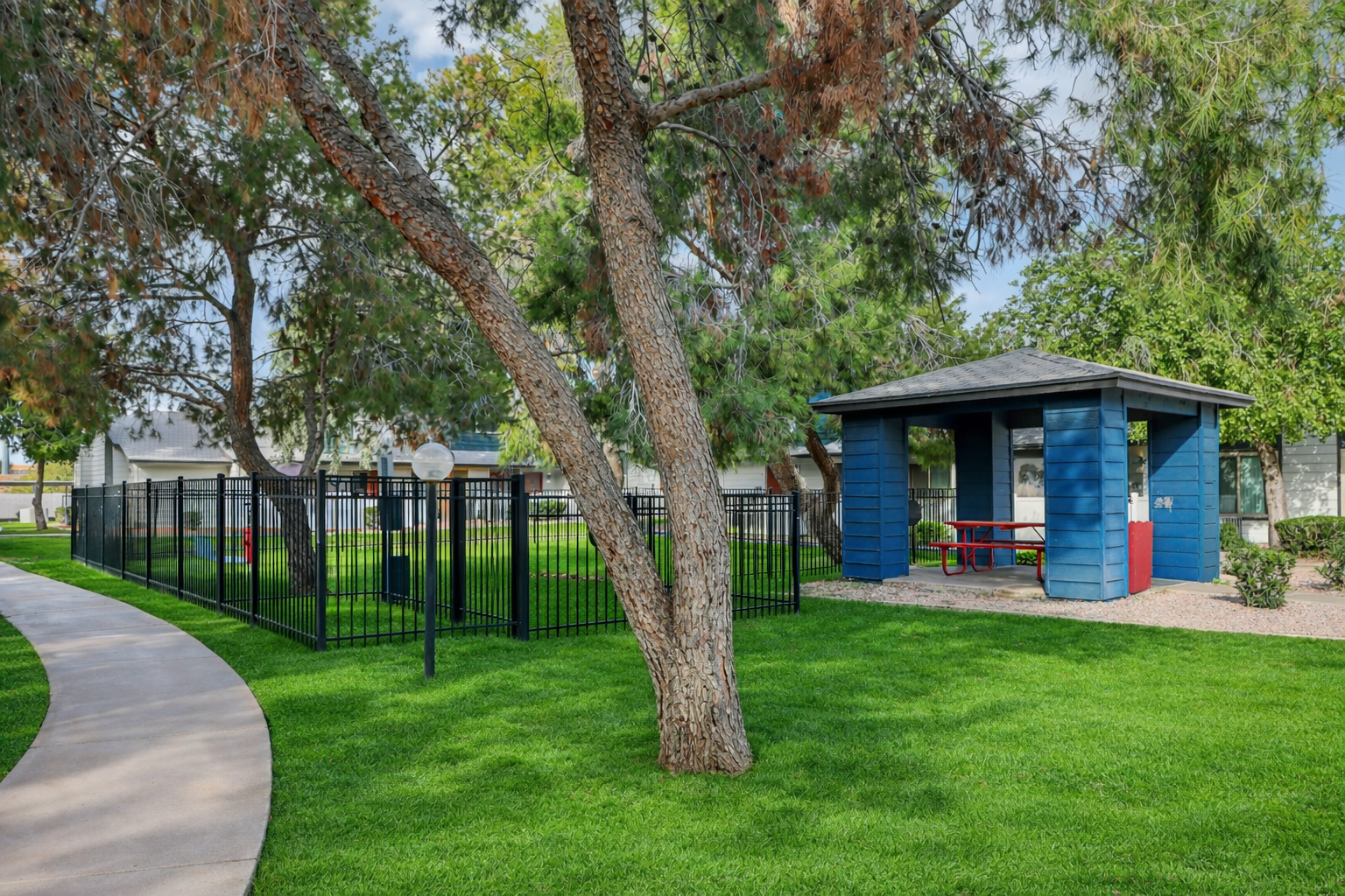 A well-maintained park featuring a curved pathway, lush green grass, and trees. There is a blue playhouse with a covered area and picnic tables, surrounded by a black fence. In the background, there are additional buildings visible, creating a pleasant outdoor space for children and families.