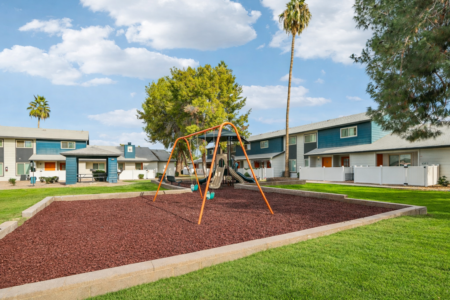 A playground with a swing set sits on a bed of red mulch, surrounded by green grass. In the background, several residential buildings are visible, featuring a mix of blue and white siding, with palm trees and a clear blue sky overhead.