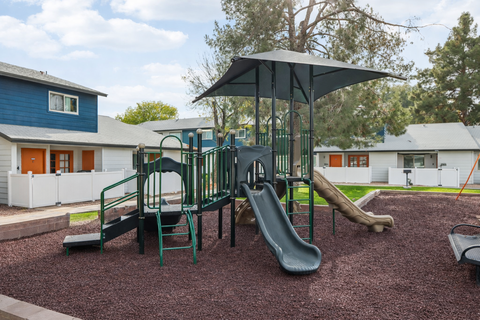 Playground featuring a multi-level structure with a slide, climbing areas, and a shaded canopy. Surrounded by mulch and residential buildings with white fences in the background under a partly cloudy sky.